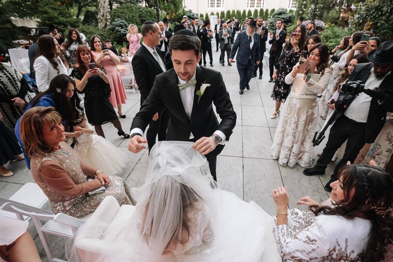 Groom bending forward smiling while guests clap during a wedding celebration outdoors along a garden walkway. Wedding at Crystal Plaza, Livingston, NJ.