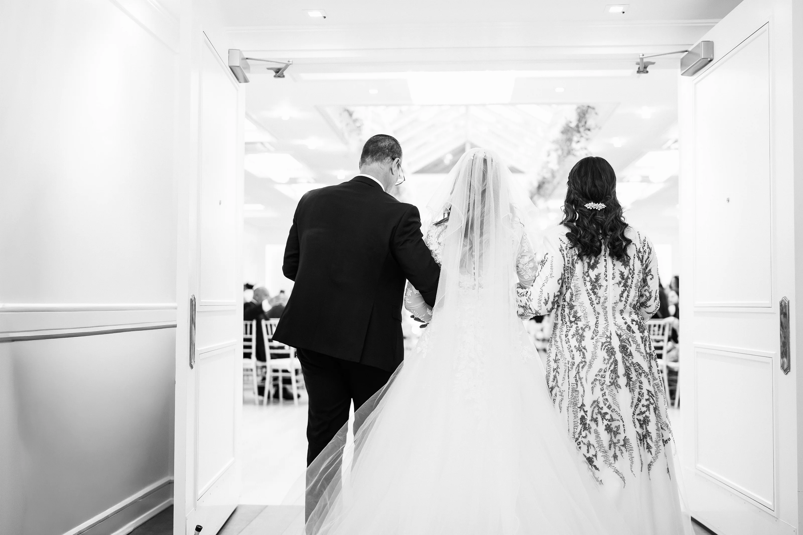 Black-and-white wedding photo of bride and groom from behind, the bride’s veil held as they walk toward the ceremony space framed by white doors.