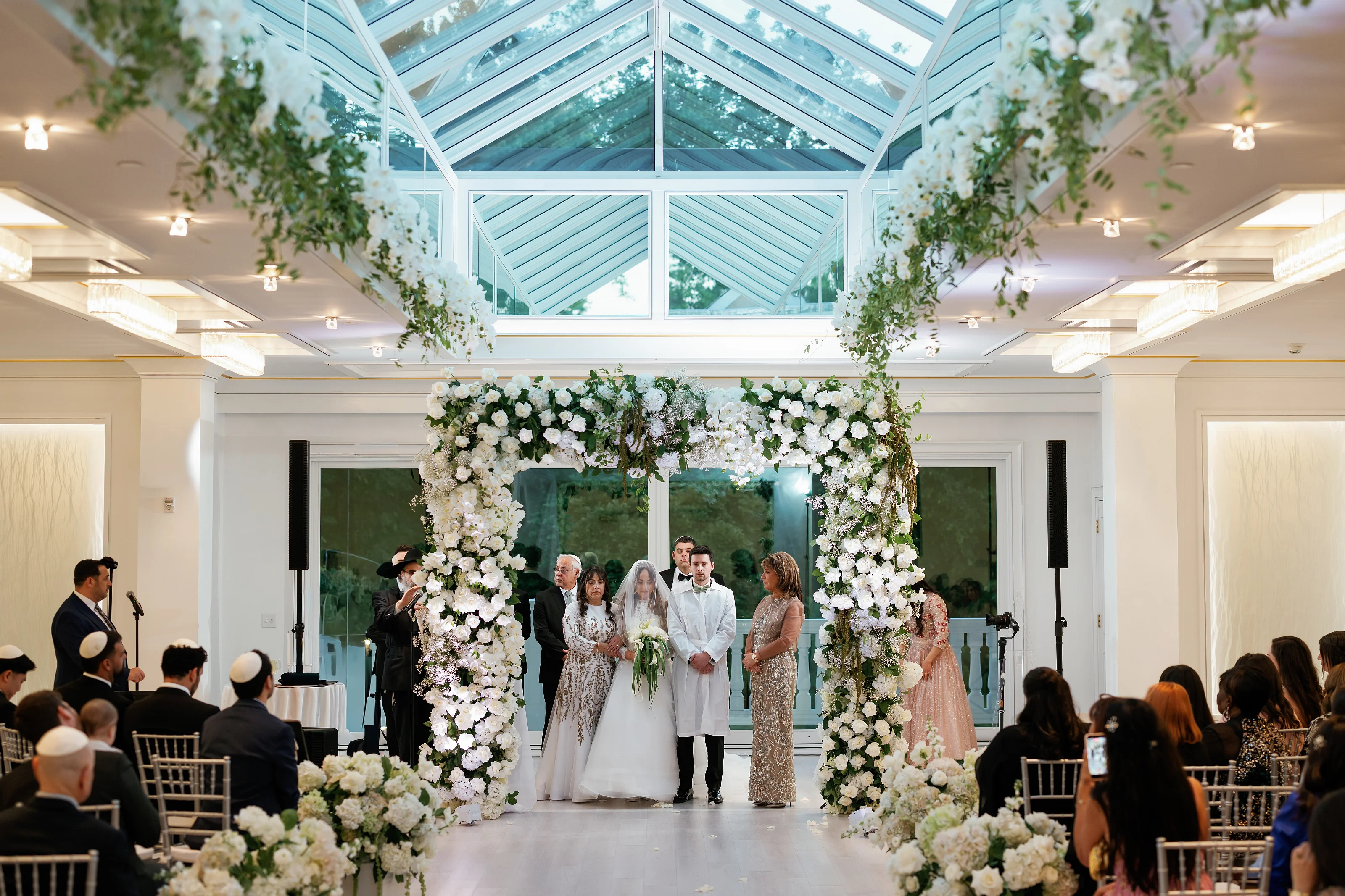 Wedding couple standing under an elaborate white floral chuppah inside a bright atrium with guests seated along the aisle.