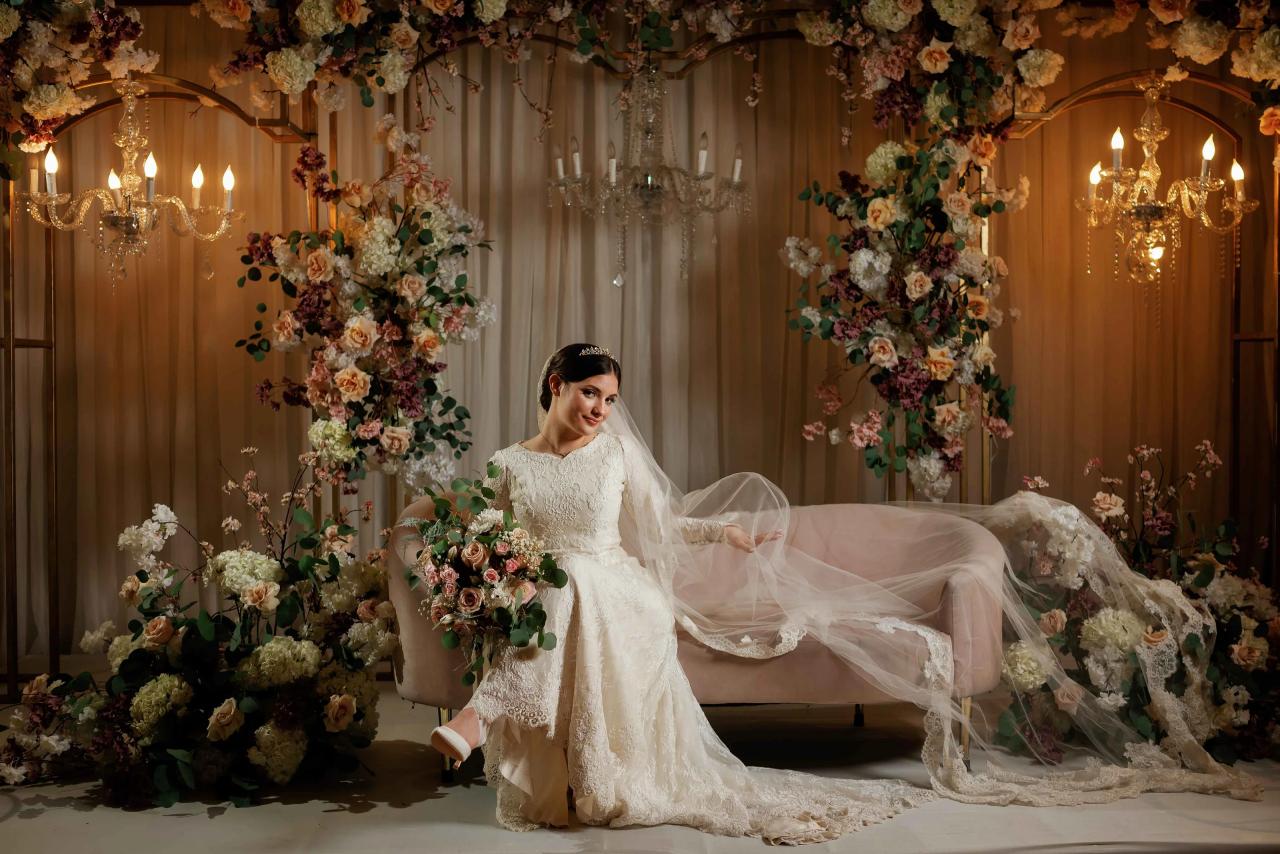 Bride in an ivory lace gown and veil reclining on a decorated chuppah-style stage, holding a bouquet of flowers under warm chandelier lighting.