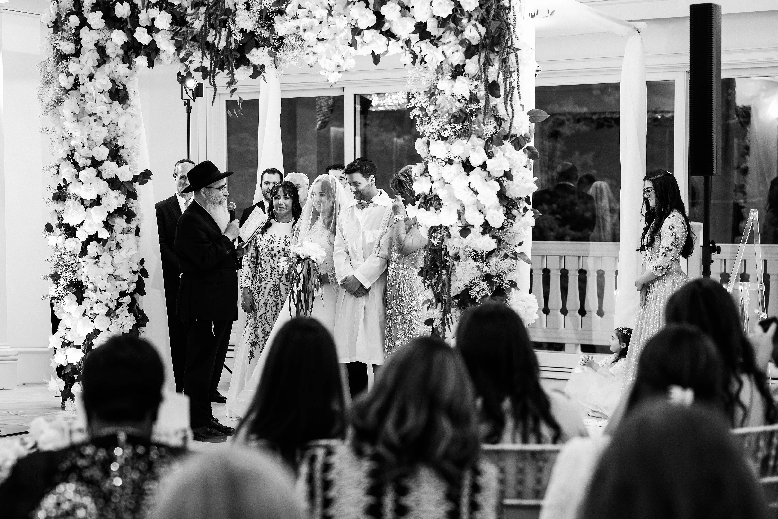 Black-and-white wedding ceremony moment during the badeken under a floral arch, with the couple centered and guests seated in the foreground.
