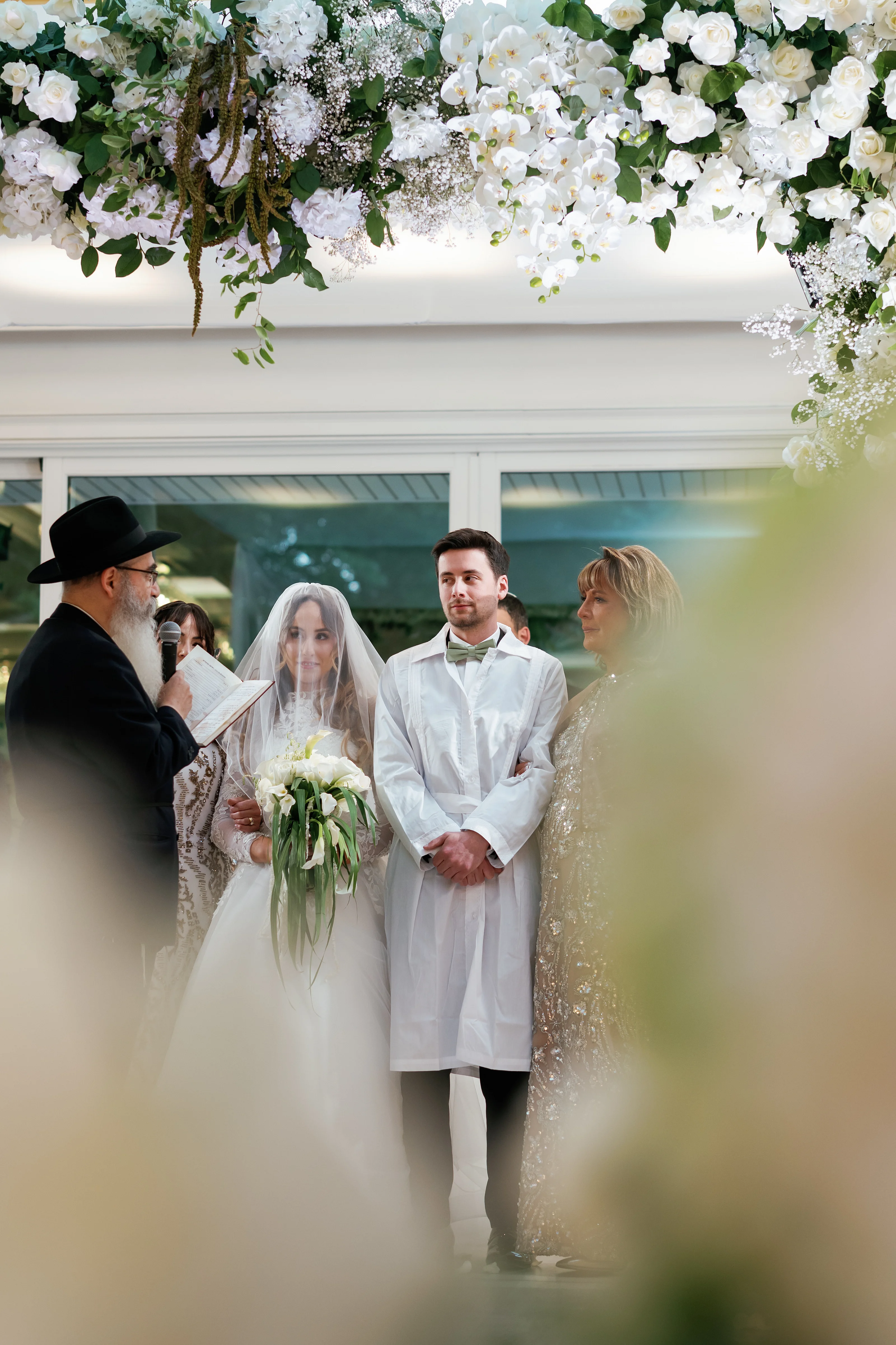 Bride and groom at the badeken moment under a floral arch with the rabbi holding a paper nearby.