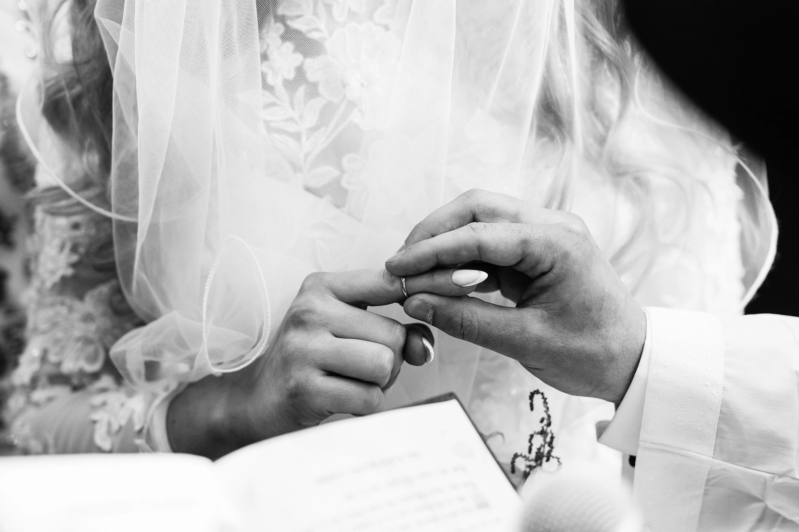 Black-and-white close-up of hands placing a wedding ring beneath a bride’s veil during a Jewish ceremony.
