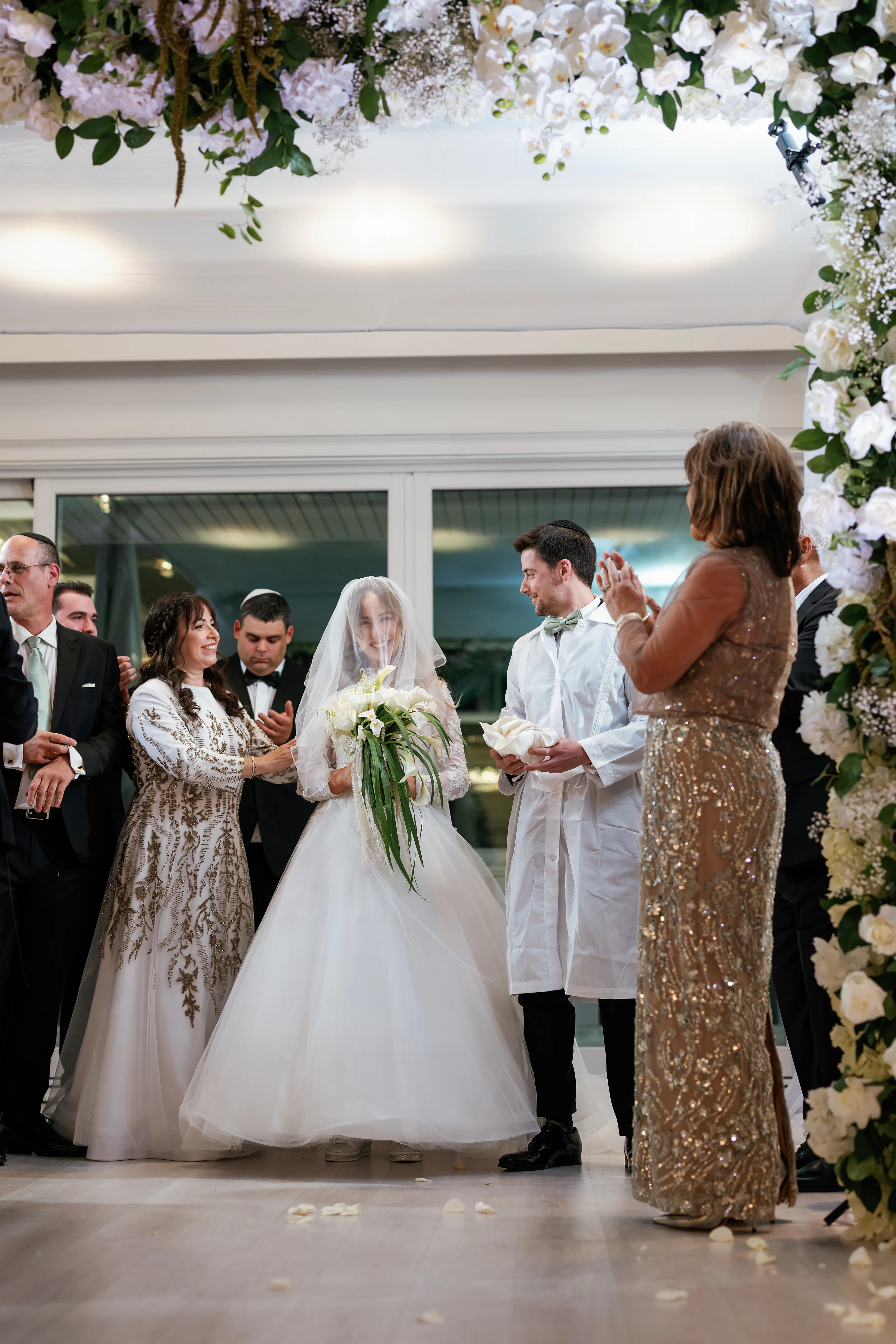 Bride and groom during the badeken moment under a floral chuppah arch, with family members nearby and soft light from large windows.