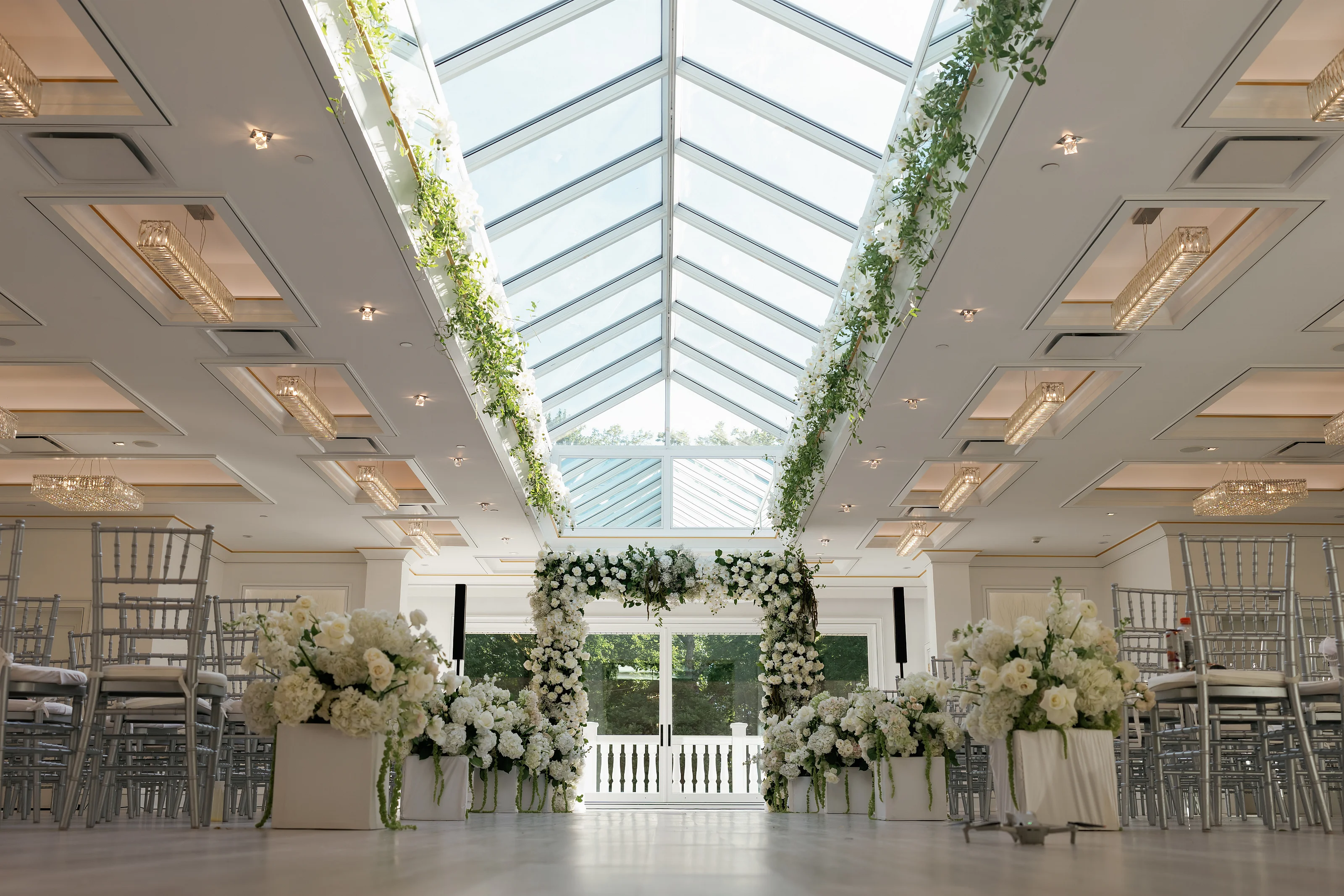 Floral chuppah ceremony setup in a bright indoor venue with tall white arrangements and greenery drapes under a vaulted glass ceiling.