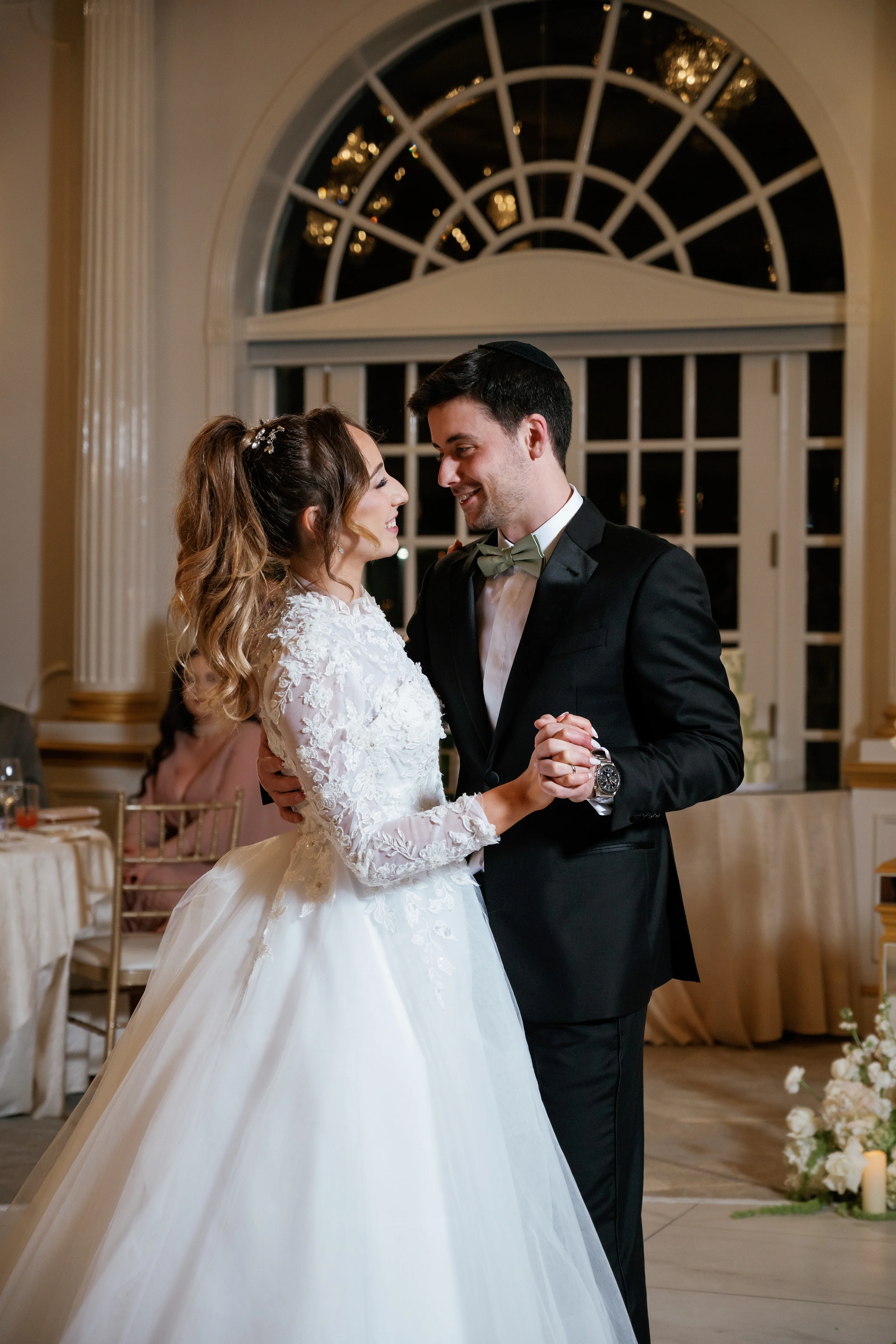 Bride and groom in a close embrace during their wedding reception, smiling at each other under warm indoor lighting with arched windows behind them.