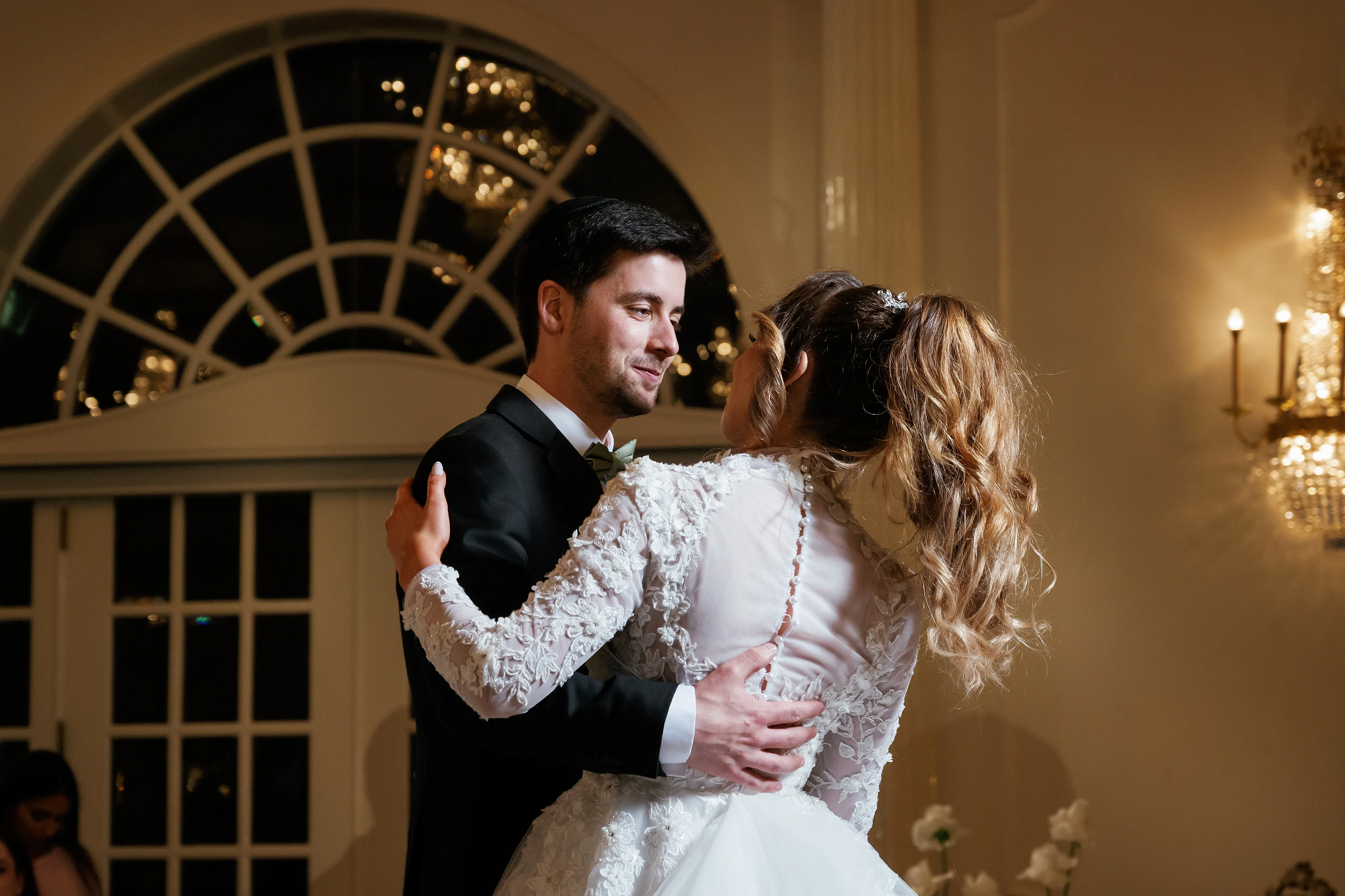 Warm indoor ceremony embrace during a Jewish wedding, with the bride’s face covered and the groom holding her close under soft golden light and arches.