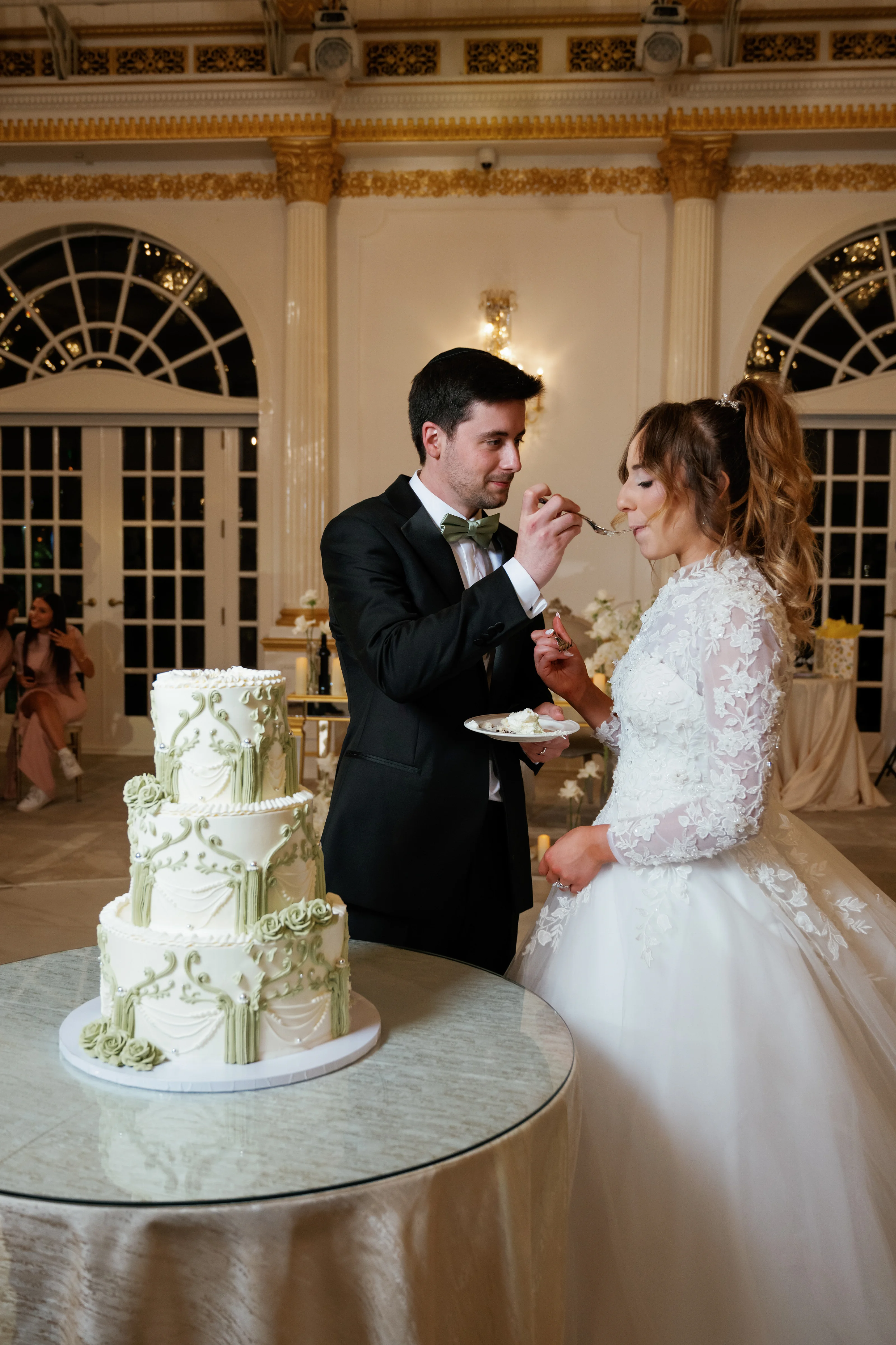 Groom and bride share a playful dessert moment, leaning in to feed each other in a warmly lit ballroom in front of a decorated wedding cake.