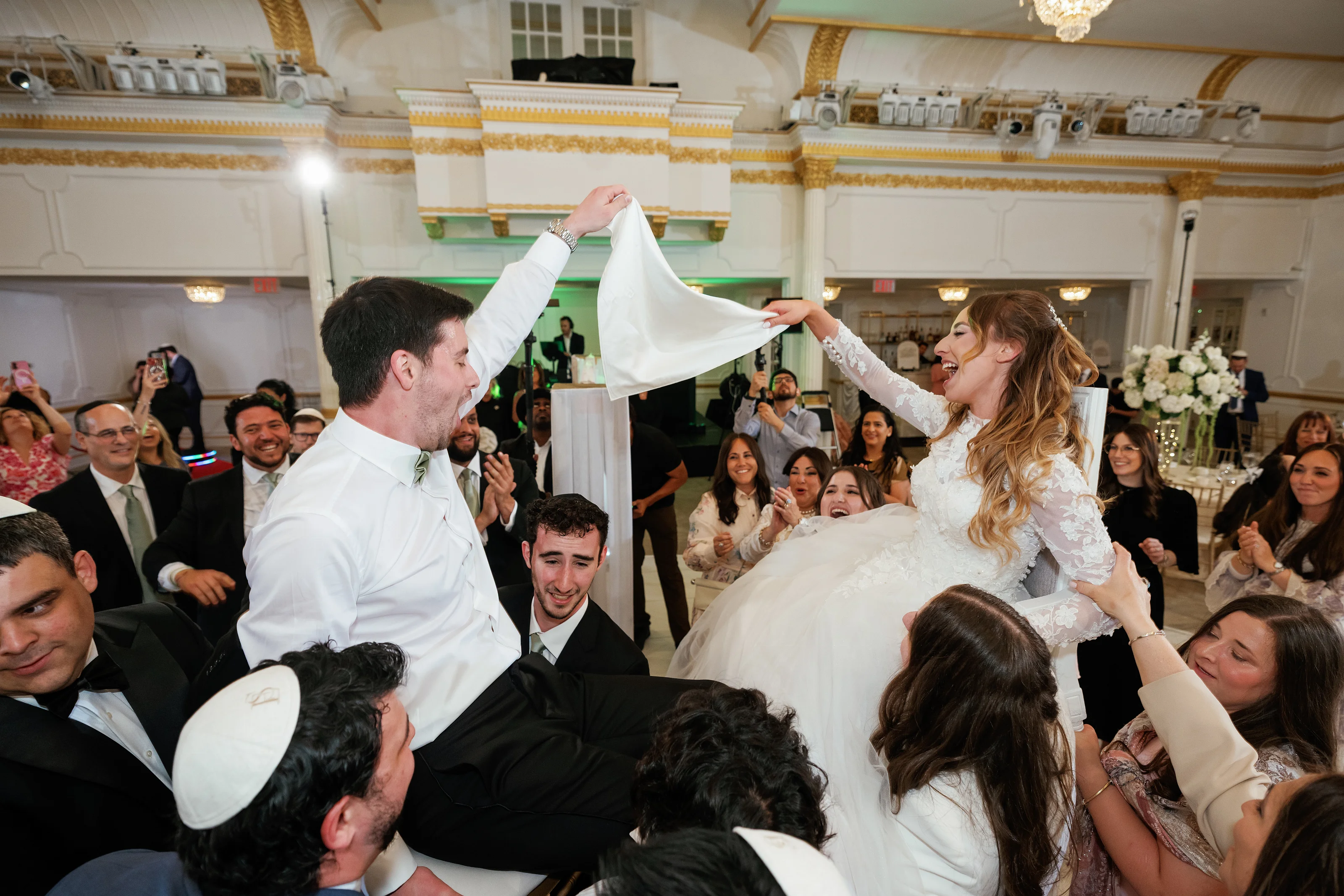 Bride and groom laughing with a white veil lifted overhead during a joyful reception finale in an ornate ballroom, surrounded by cheering guests.