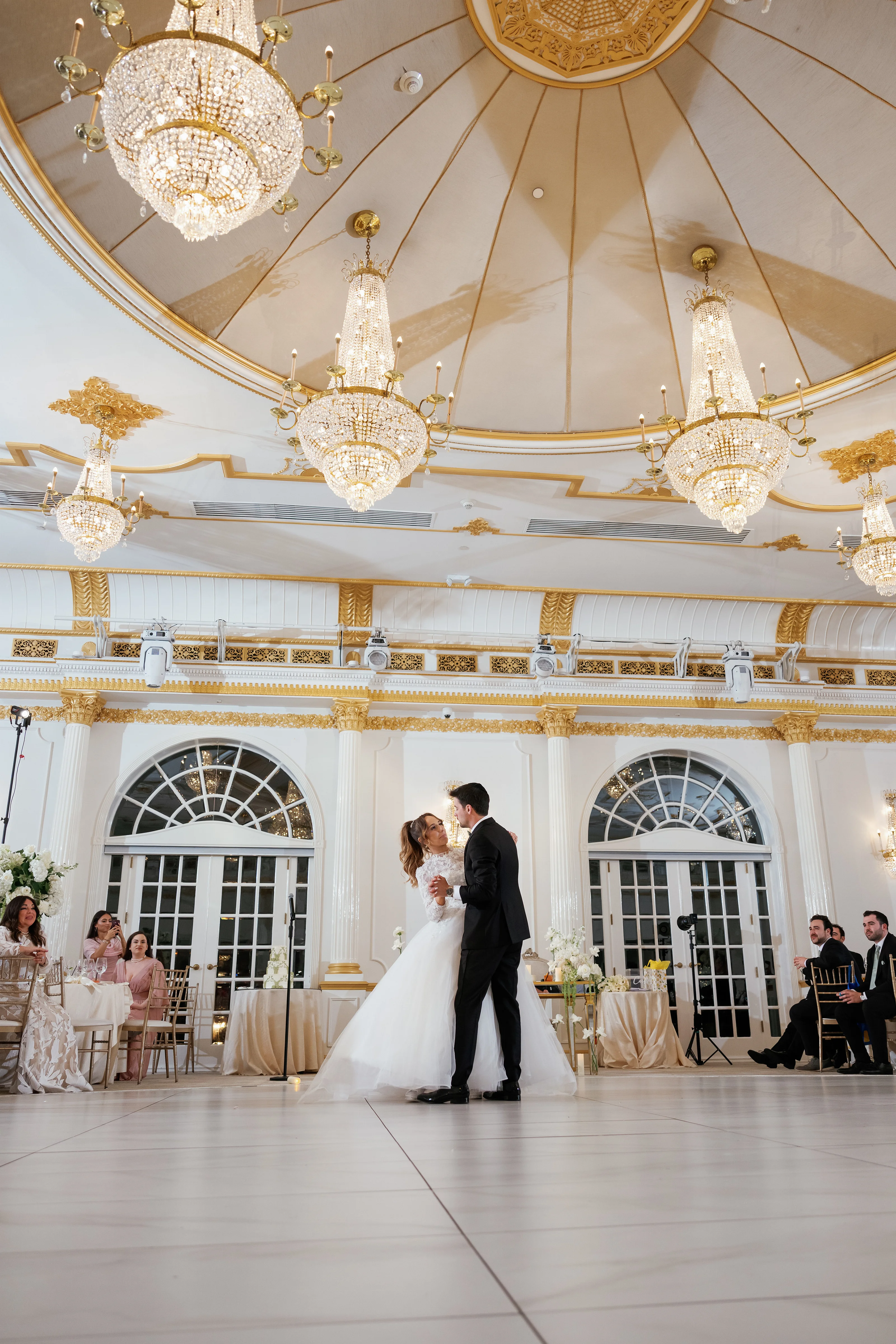 Wedding couple standing close and facing each other in a grand indoor ceremony hall beneath ornate gold and crystal chandeliers.