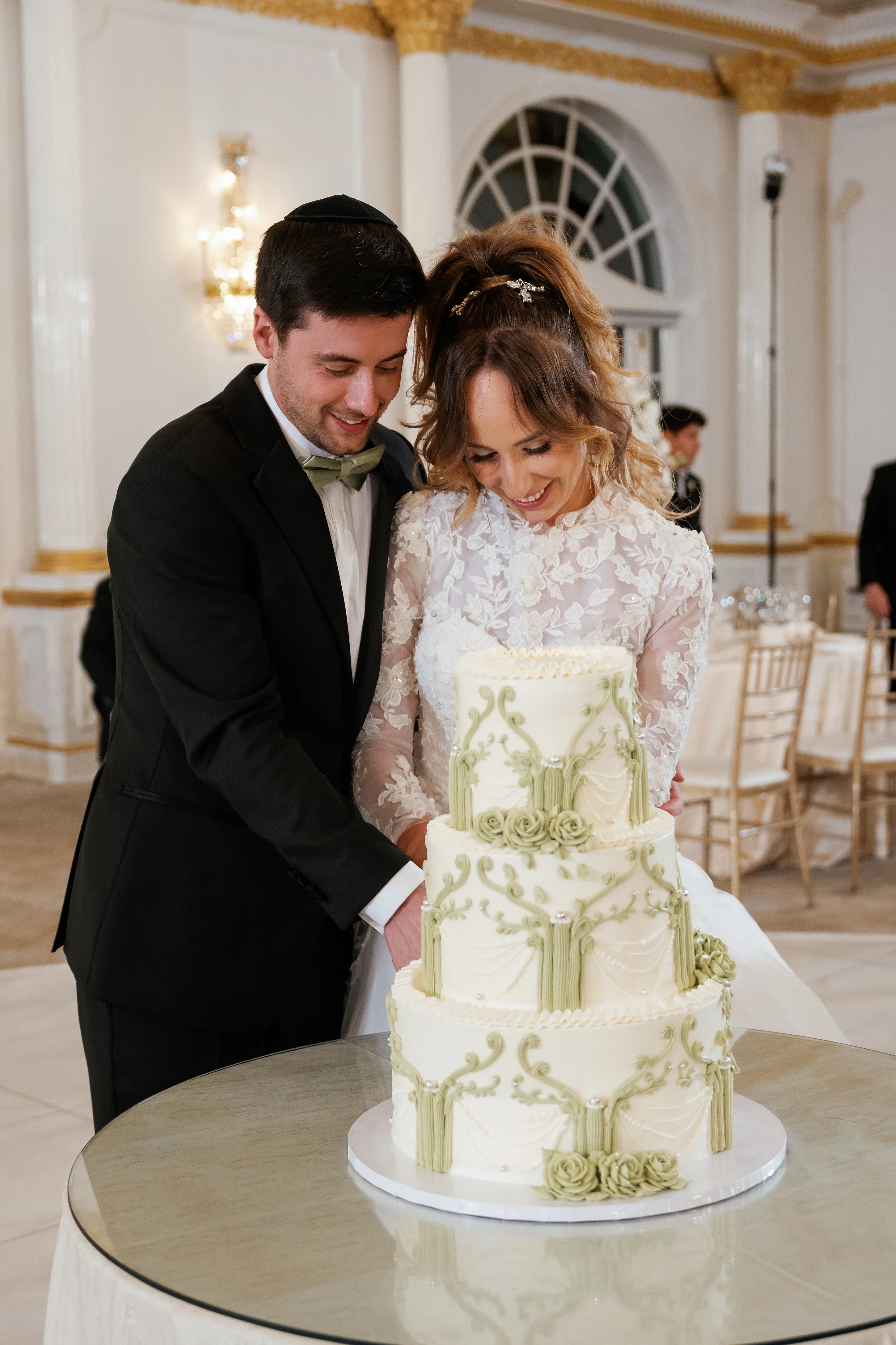 Bride and groom lean in together to cut a decorated wedding cake during the reception, captured in warm indoor lighting with an elegant hall backdrop.