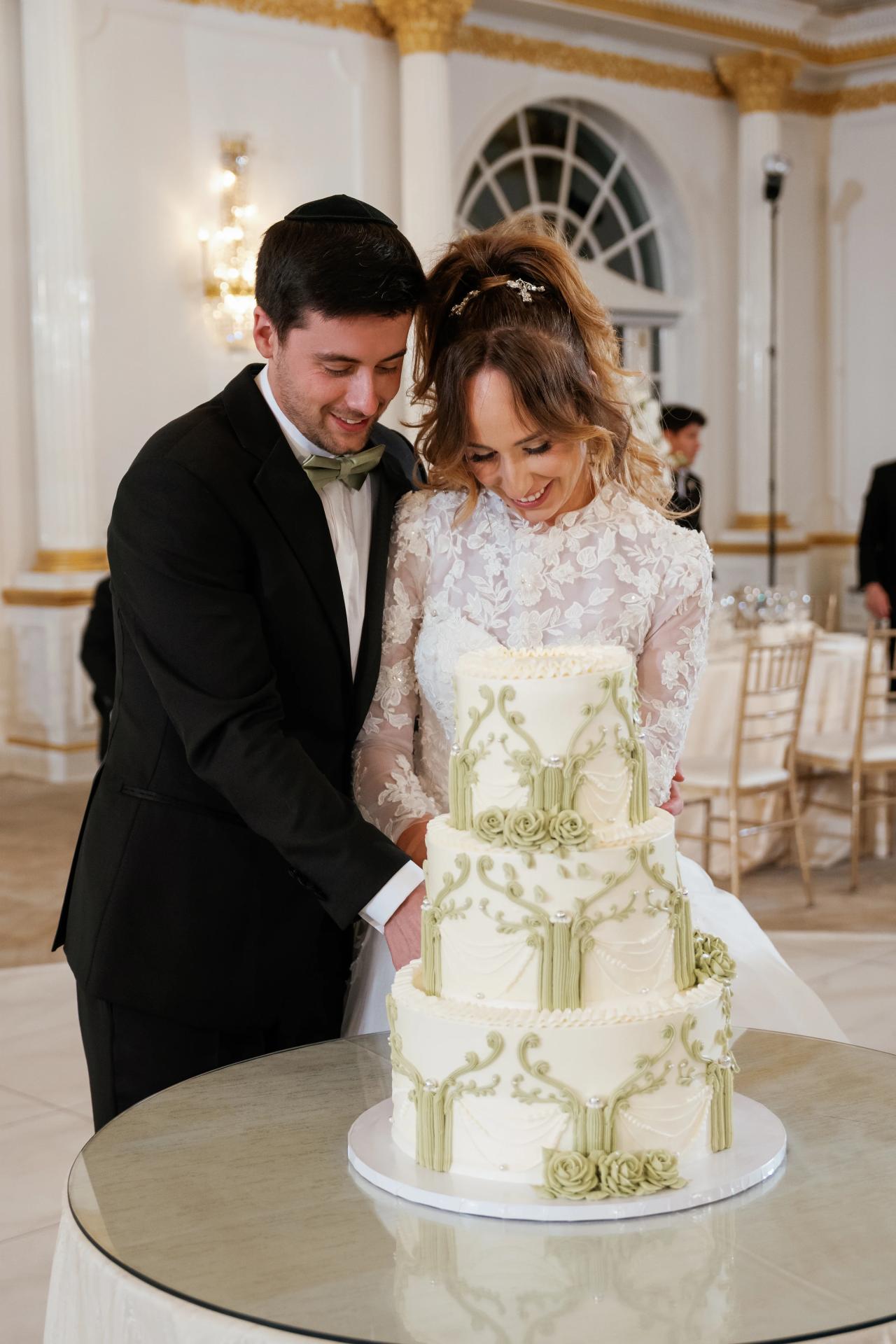 Bride and groom lean in together to cut a decorated wedding cake during the reception, captured in warm indoor lighting with an elegant hall backdrop. Wedding at Crystal Plaza, Livingston, NJ.