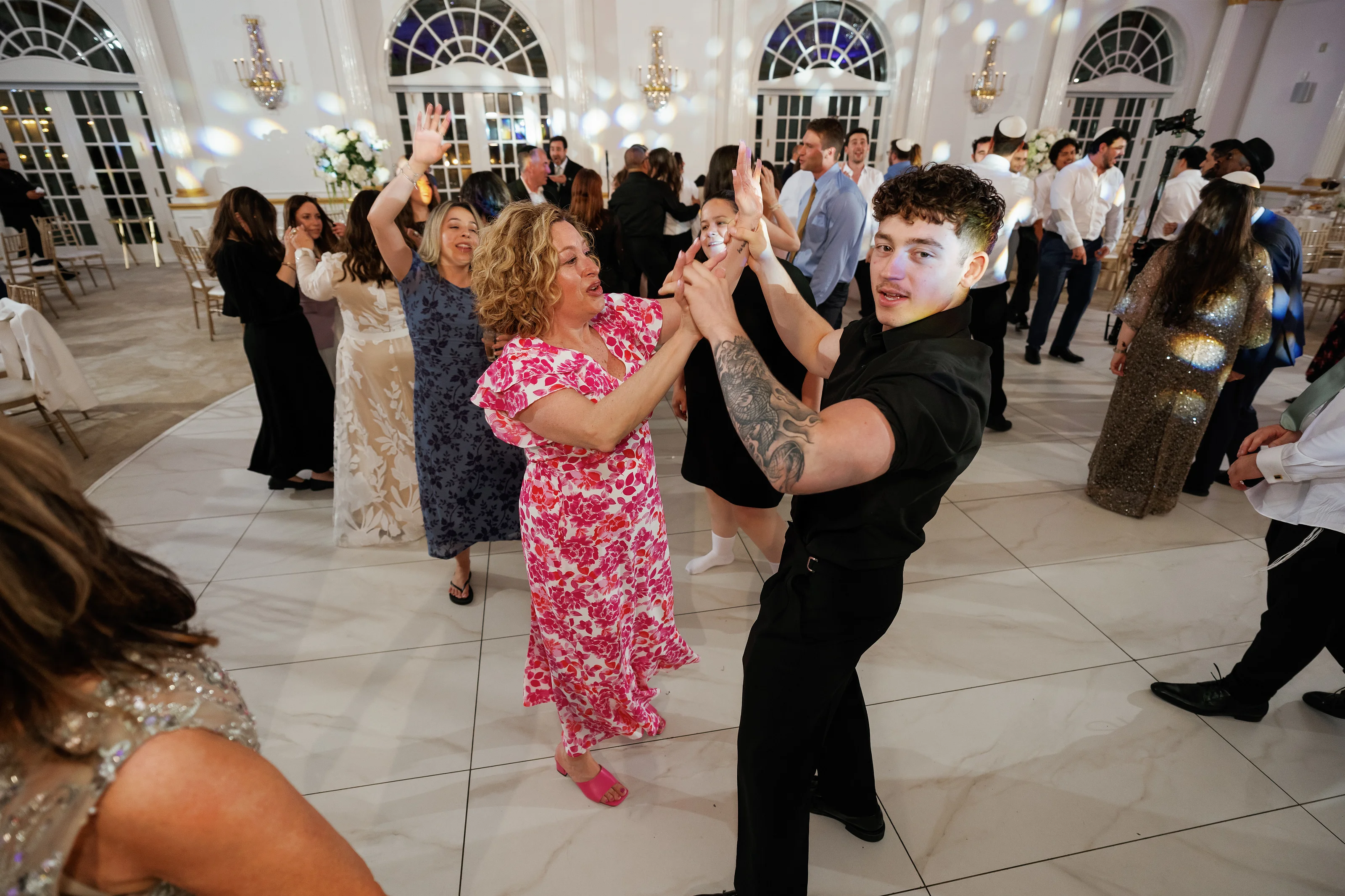 Wedding couple dancing joyfully in a bright ballroom with guests celebrating in the background under chandeliers.