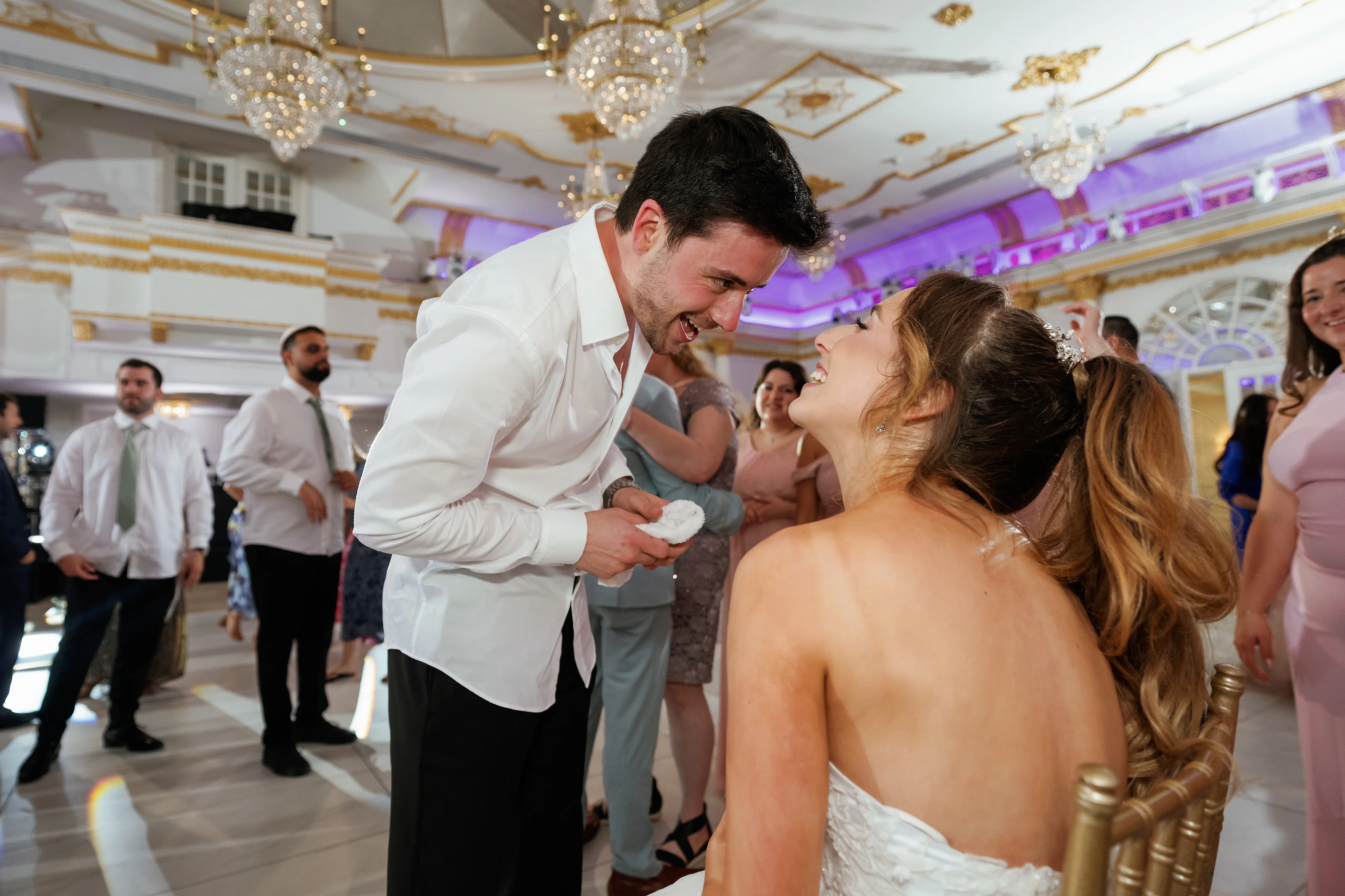Groom and bride share a joyful laugh in a chandelier-lit ballroom during the celebration as guests watch in the background.
