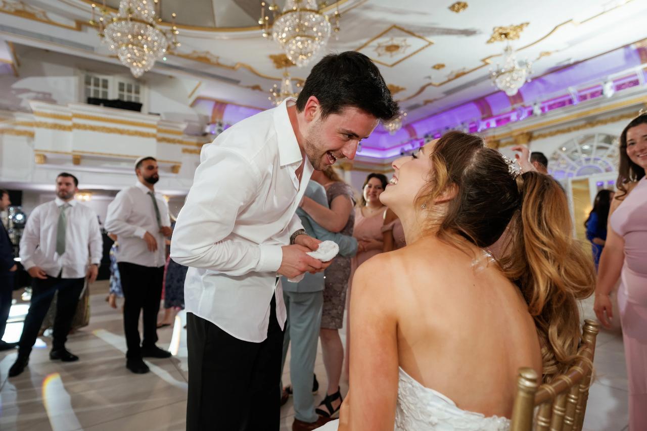 Groom and bride share a joyful laugh in a chandelier-lit ballroom during the celebration as guests watch in the background. Wedding at Crystal Plaza, Livingston, NJ.