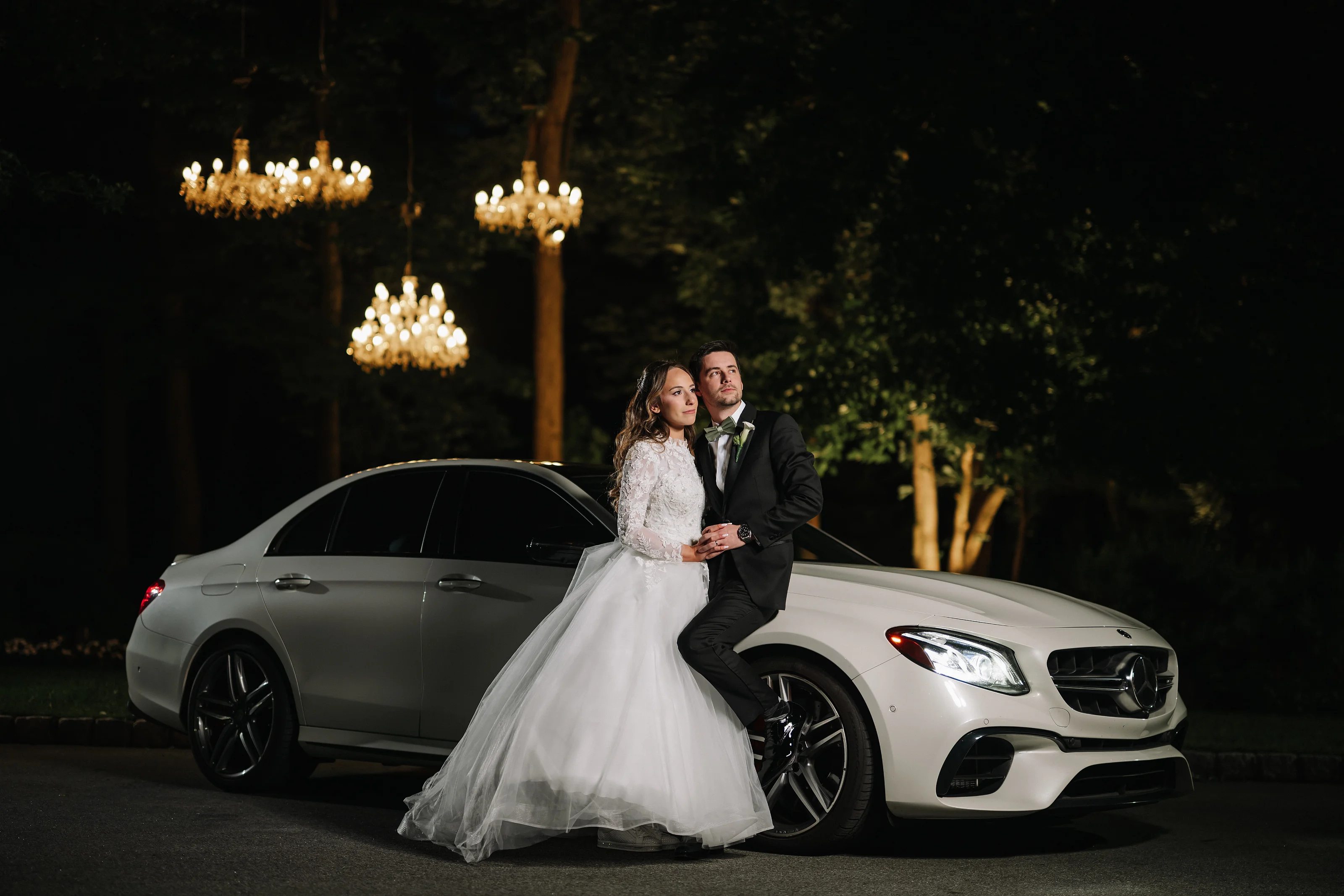 Bride and groom standing in an intimate embrace beside a white sports car at night, lit by warm chandelier-style lights outdoors.