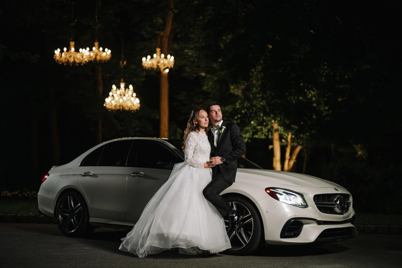 Bride and groom standing in an intimate embrace beside a white sports car at night, lit by warm chandelier-style lights outdoors.