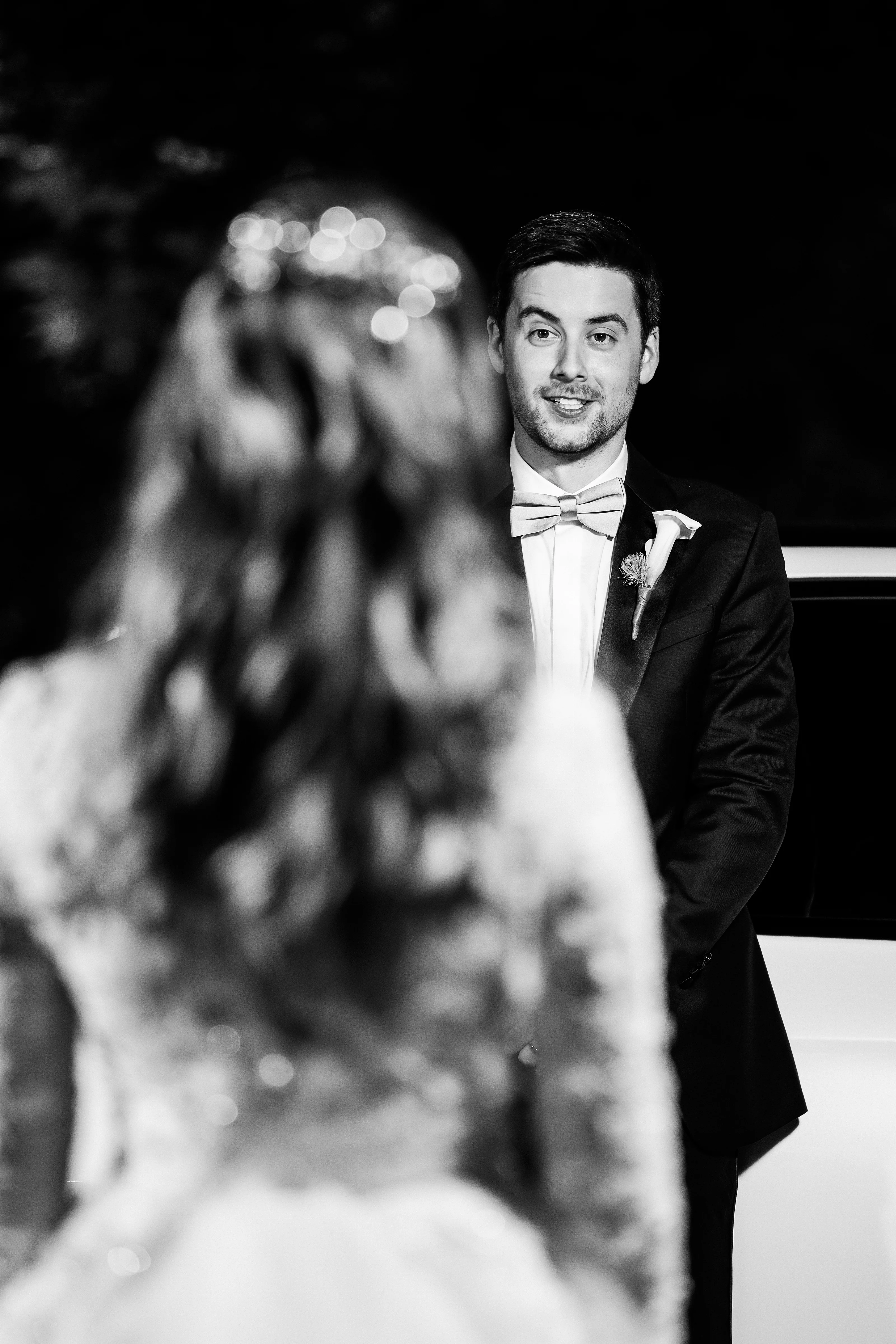 Black-and-white portrait of a groom in a tuxedo with bow tie during the veil moment, with the bride’s out-of-focus veil in the foreground and soft bokeh lights behind.