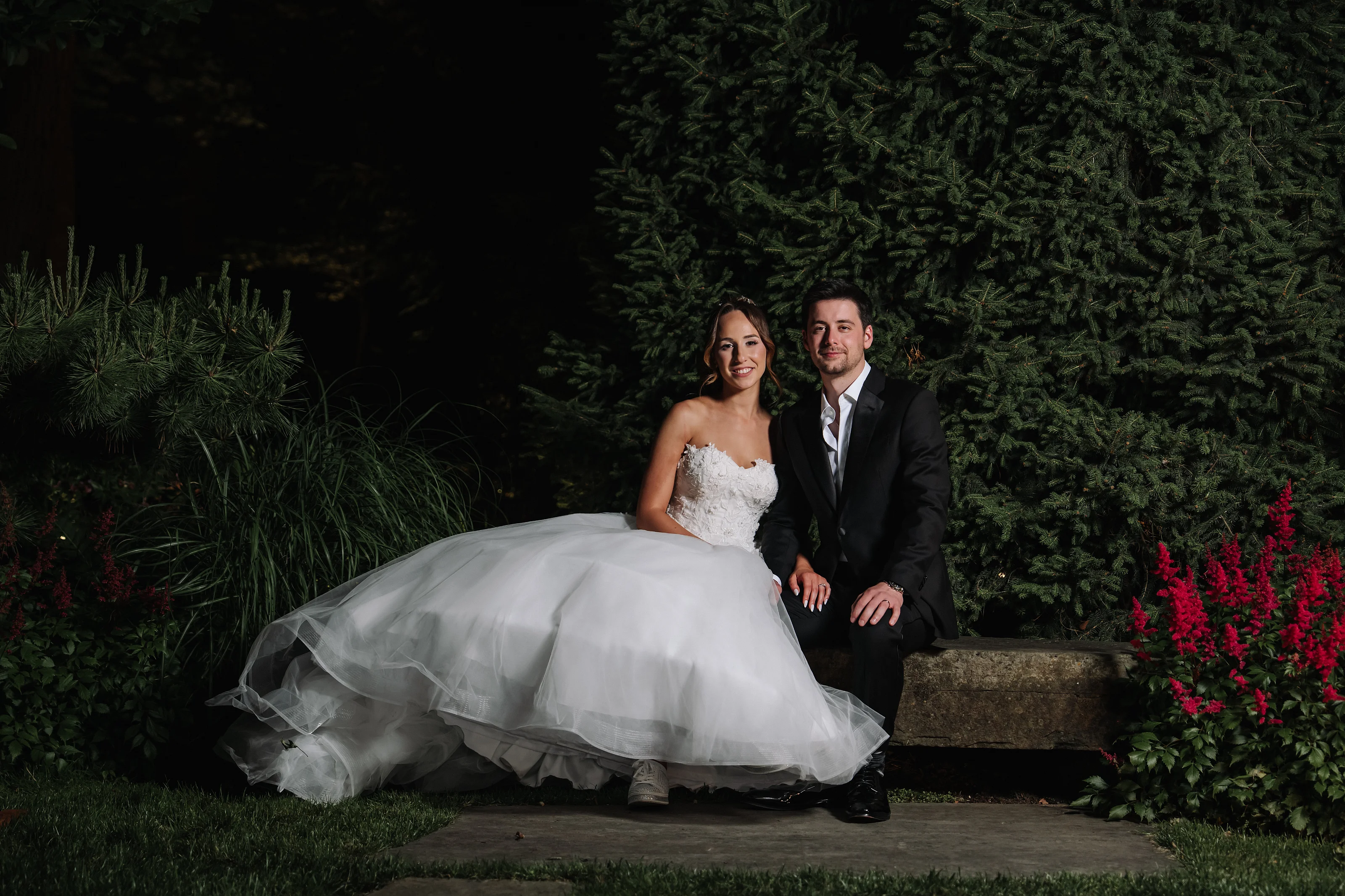 Bride and groom seated together on a stone ledge in a landscaped garden at night, with greenery and red flowers in the background.