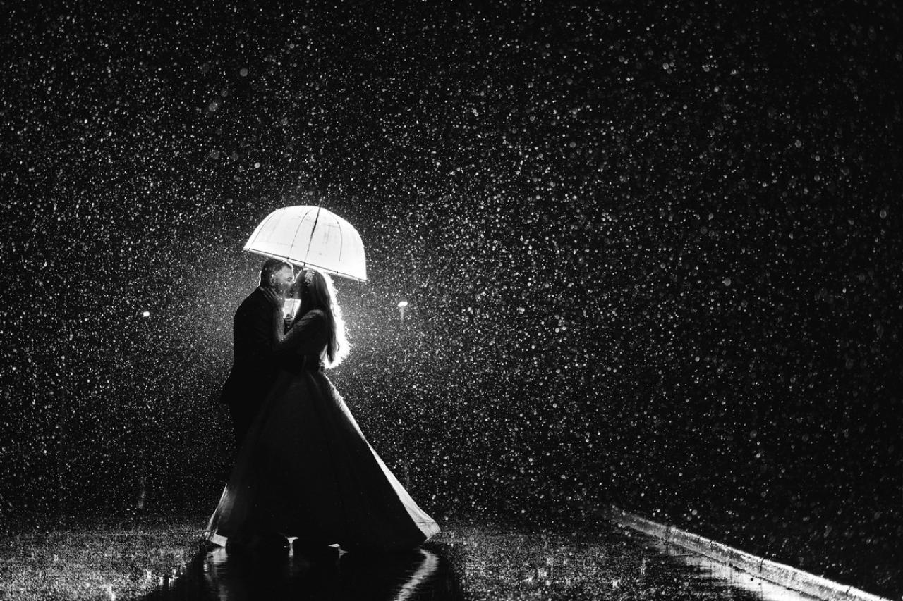Black-and-white photo of a couple embracing under an open umbrella in the rain, with water droplets sparkling in the air.