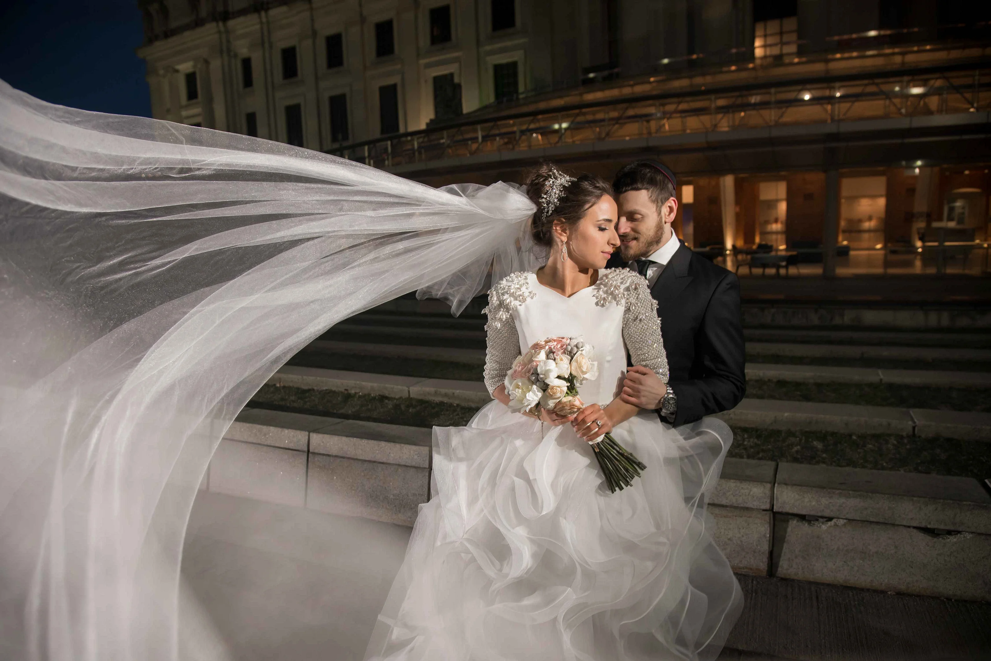 Bride in a white gown holding a bouquet while her veil sweeps across the frame; groom in a black suit embraces her at night outside the venue.