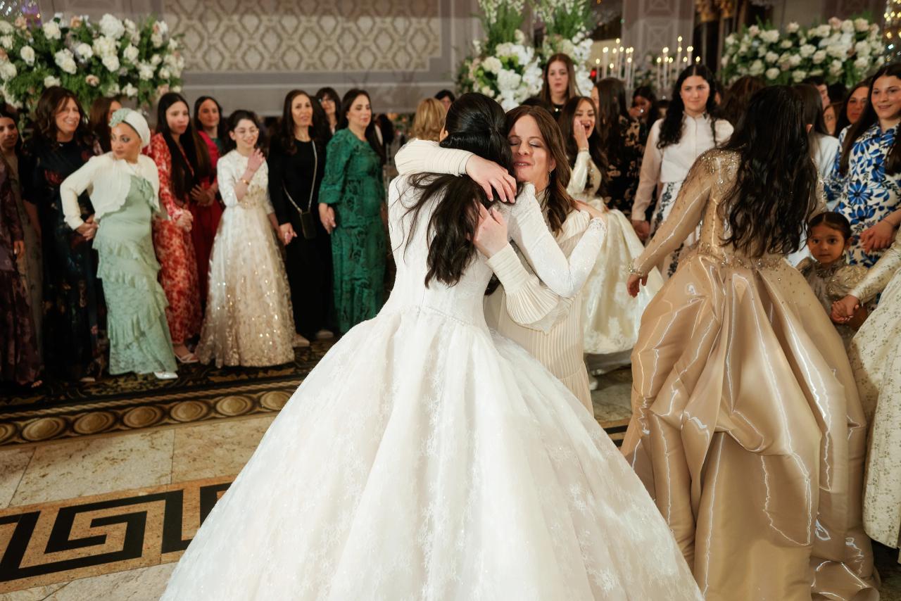 Bride and groom embrace in the middle of a lively wedding celebration as the crowd looks on in a warmly lit hall with floral décor.
