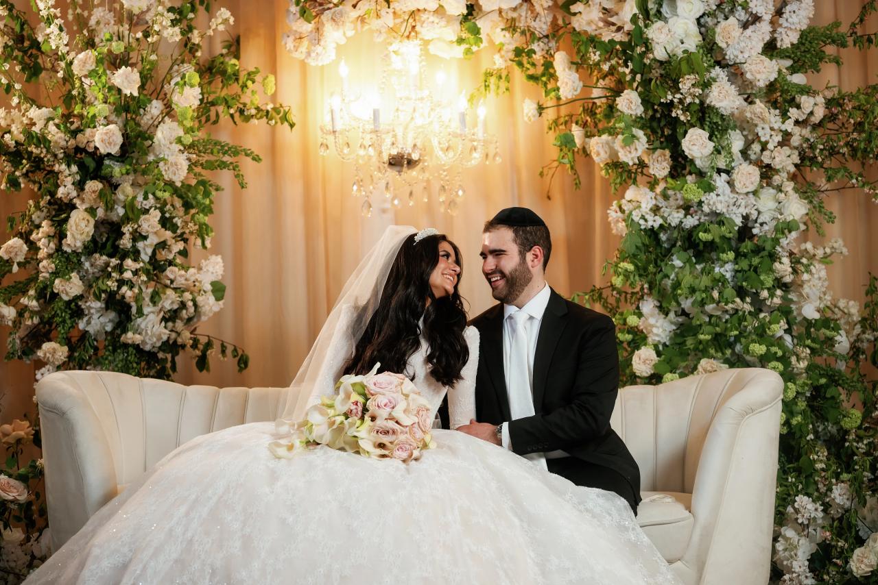 Bride and groom seated together smiling at each other during a floral-filled ceremony under warm chandelier light.