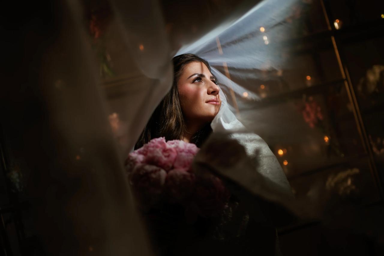 Bride in a sheer veil moment, gazing upward with warm indoor lights in the background, framed by fabric for an intimate cinematic look.