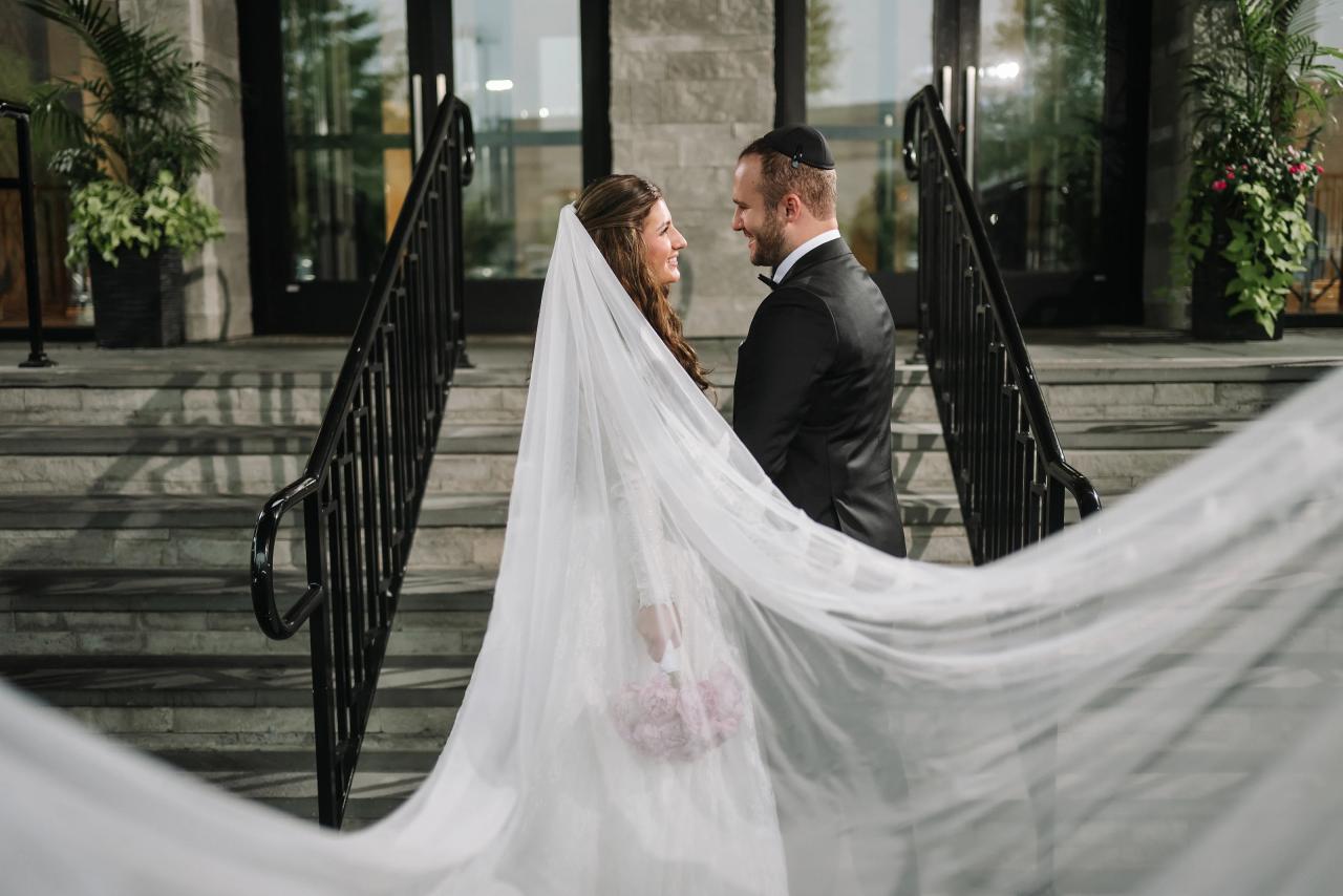 Bride and groom share an intimate veil moment on stone steps framed by black railings, with warm ambient light and flowing veil fabric.