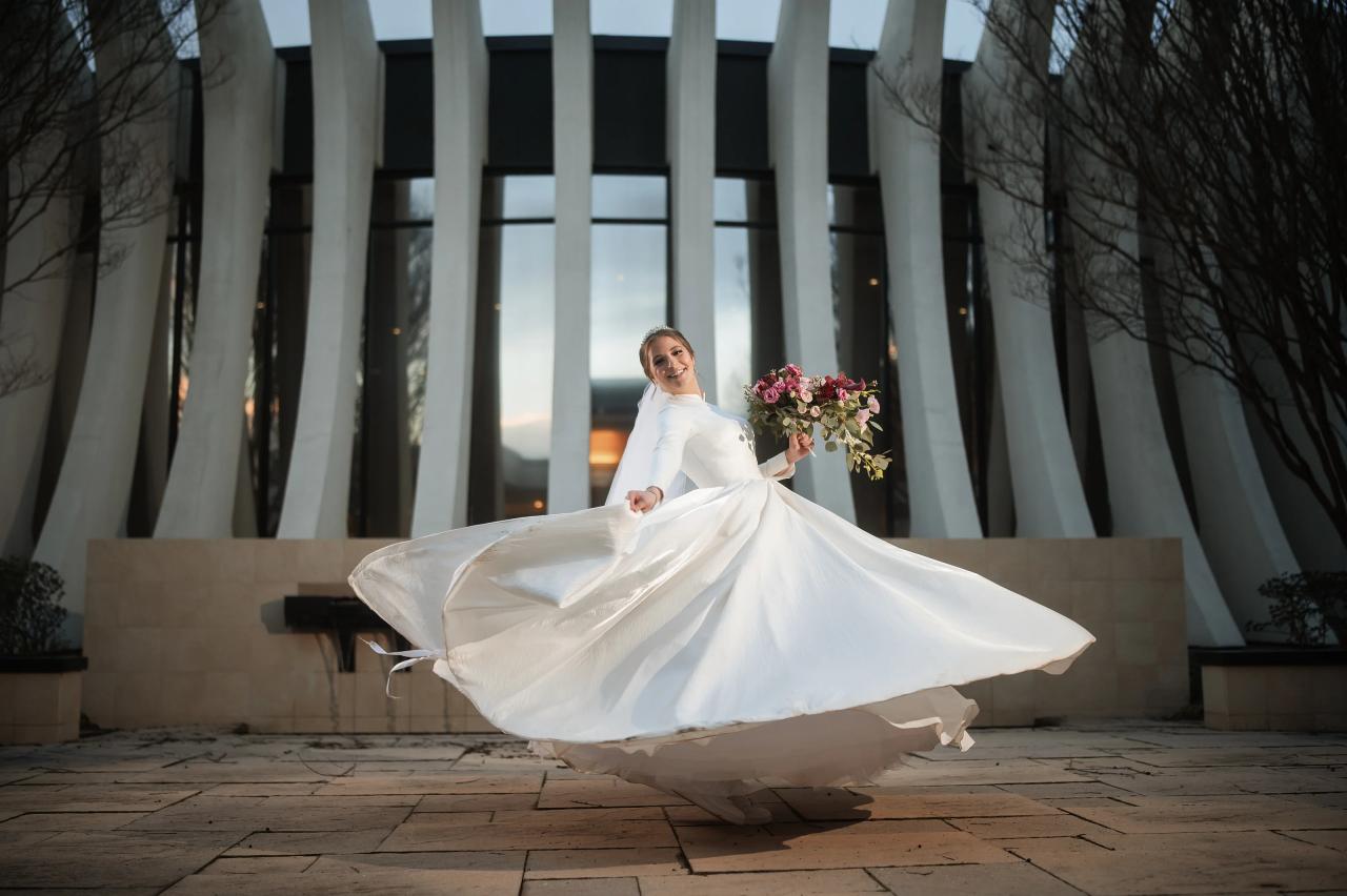 Bride in a white dress holding a bouquet, spinning with her veil flowing in a courtyard with tall columns at sunset.