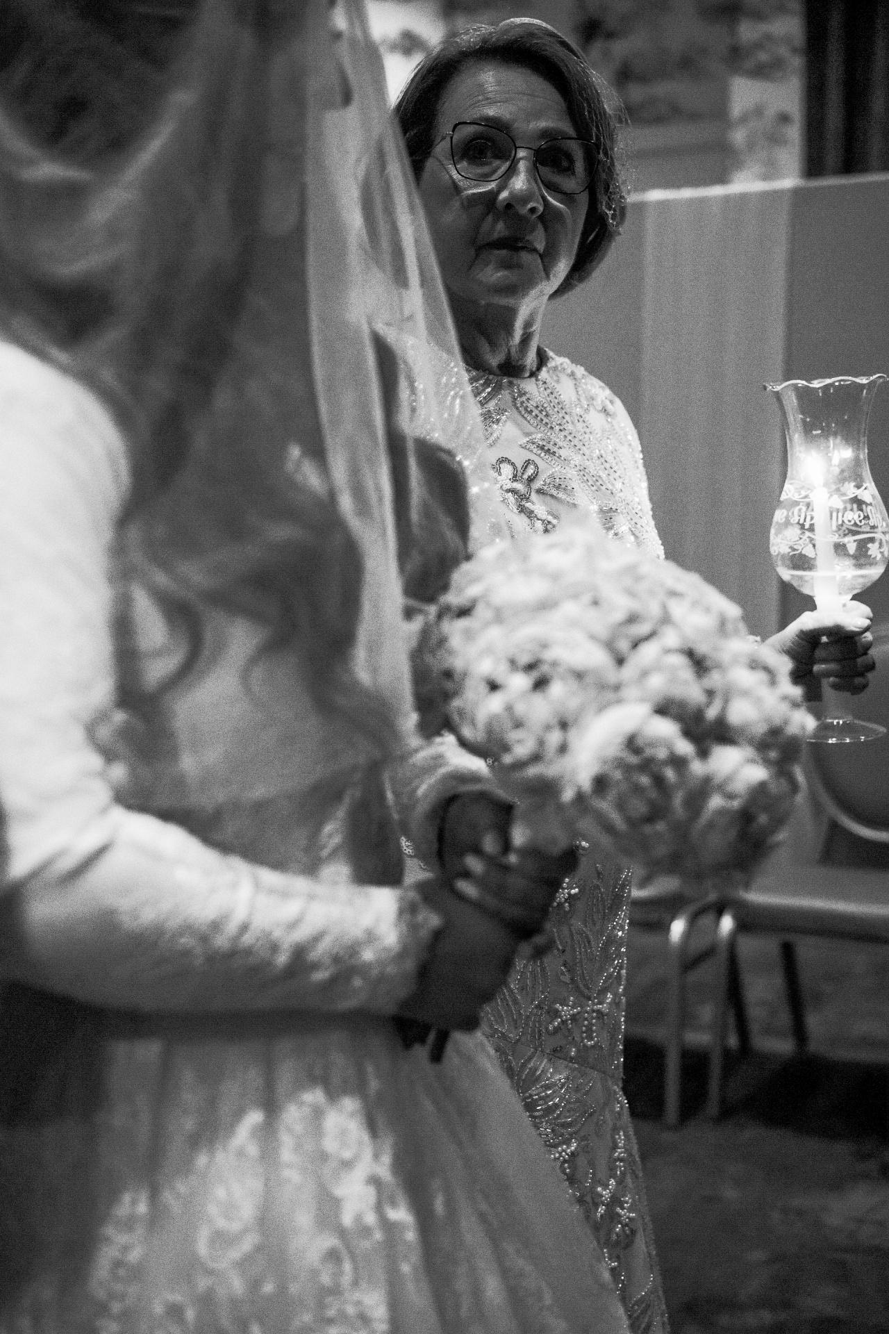 Black-and-white candid of an older woman holding a drink at a celebration, facing the camera with an emotional expression while another guest stands in the foreground.