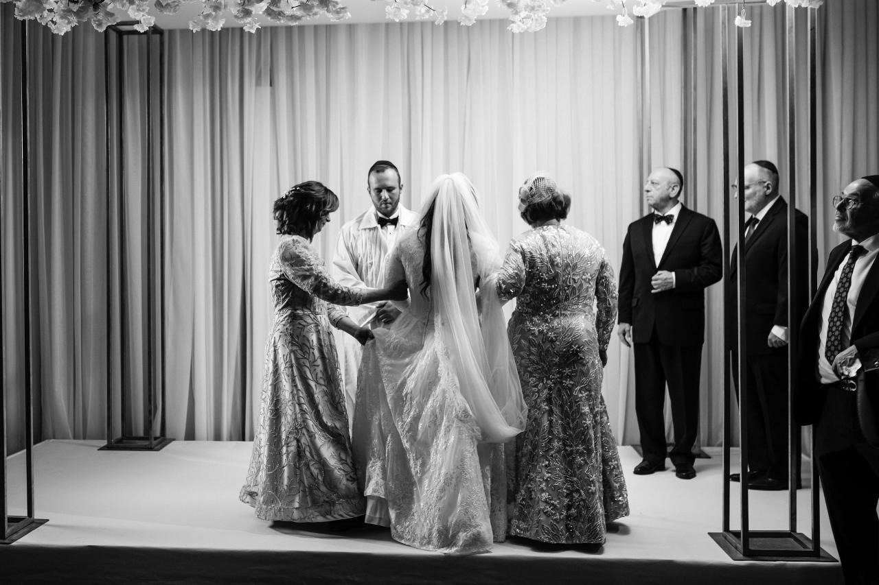 Black-and-white ceremony stage photo showing a bride and groom during the badeken moment with family members standing nearby, draped curtains in the background.