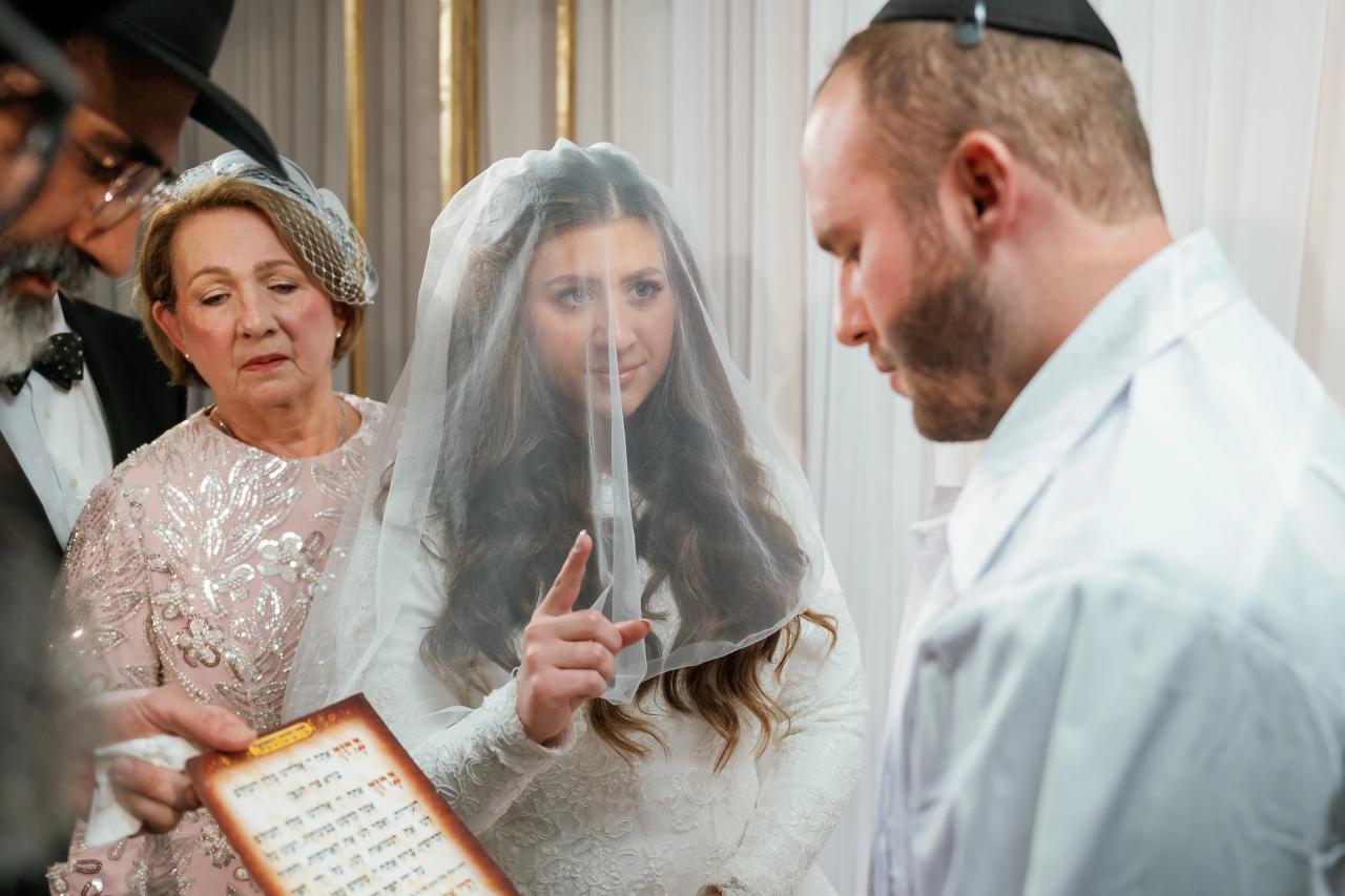 Bride in a white lace dress holding her veil forward with her fingers during badeken, with the groom in profile nearby and family members behind her.