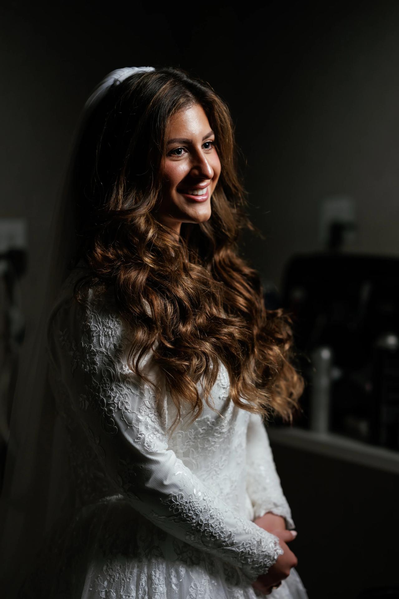 Bride portrait with long wavy hair and lace dress, smiling softly in dramatic low-key lighting against a dark background.