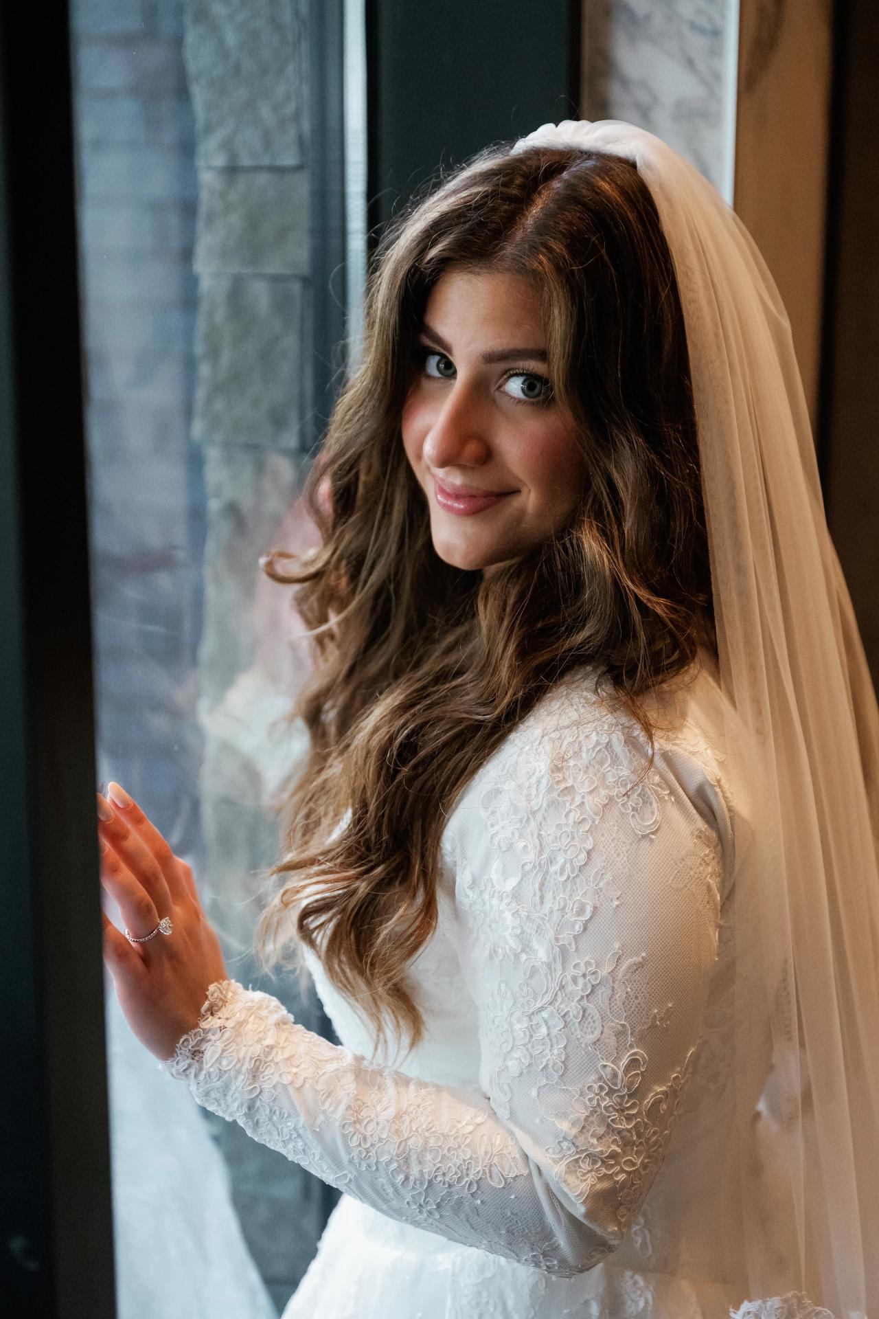 Close portrait of a bride in a white lace dress and veil, smiling gently and raising one hand indoors against a stone wall background.