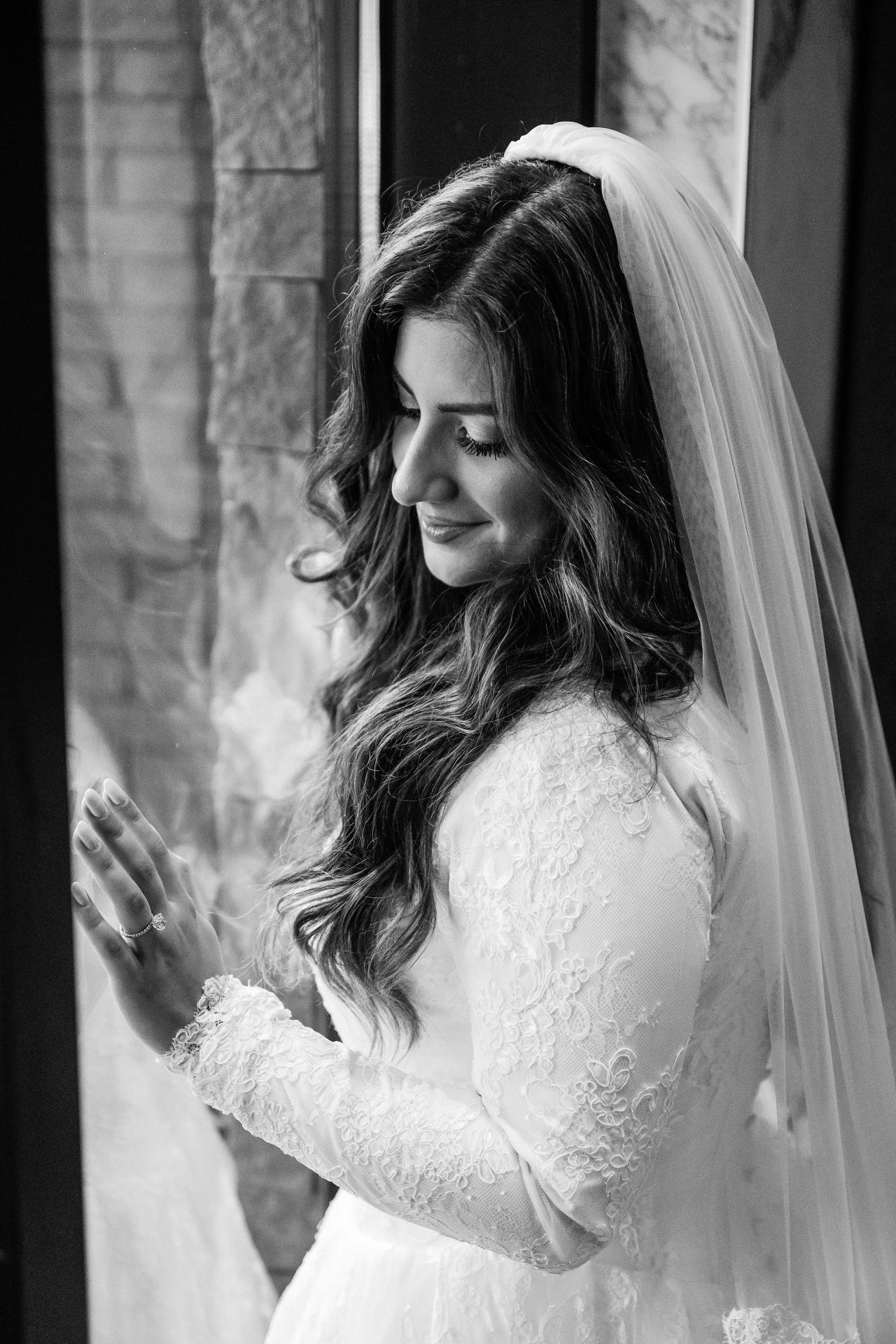 Black-and-white portrait of a bridal bride in a veil with eyes closed and hand raised, lit softly indoors against a stone wall.