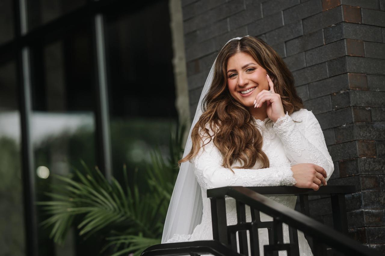 Brides portrait smiling while leaning on a dark balcony railing with a veil draped over her shoulders against a stone wall.