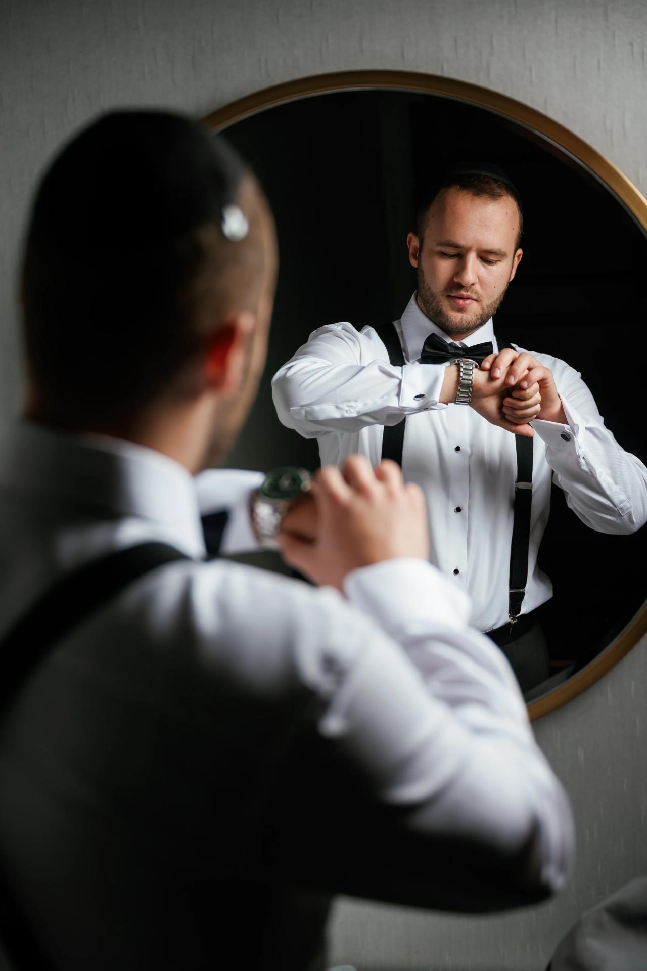 Groom adjusting his wristwatch while getting ready, seen through a mirror with soft indoor lighting.