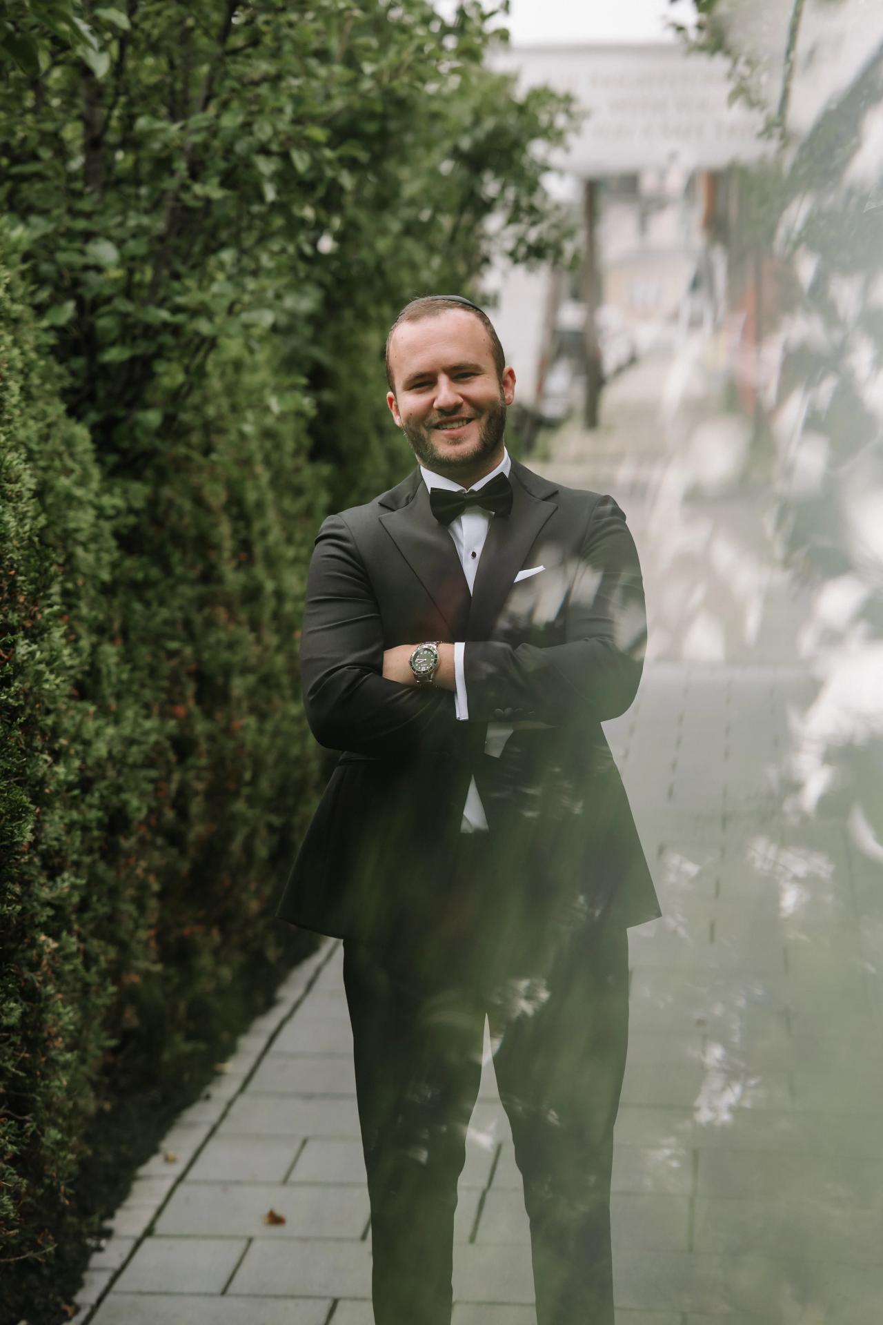 Smiling groom in a dark suit with bow tie posed with arms crossed in a lush garden courtyard, soft bright light and shallow depth of field.