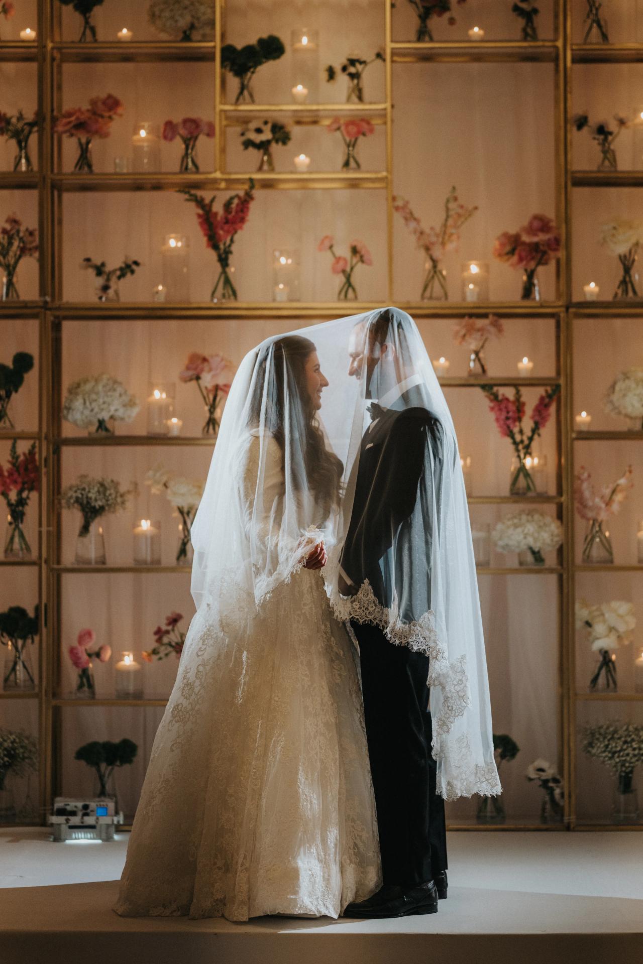 Bride and groom share a close moment under the bride’s veil during the badeken, warmly lit in an indoor ceremony space with floral decor and candle-like lights behind them.