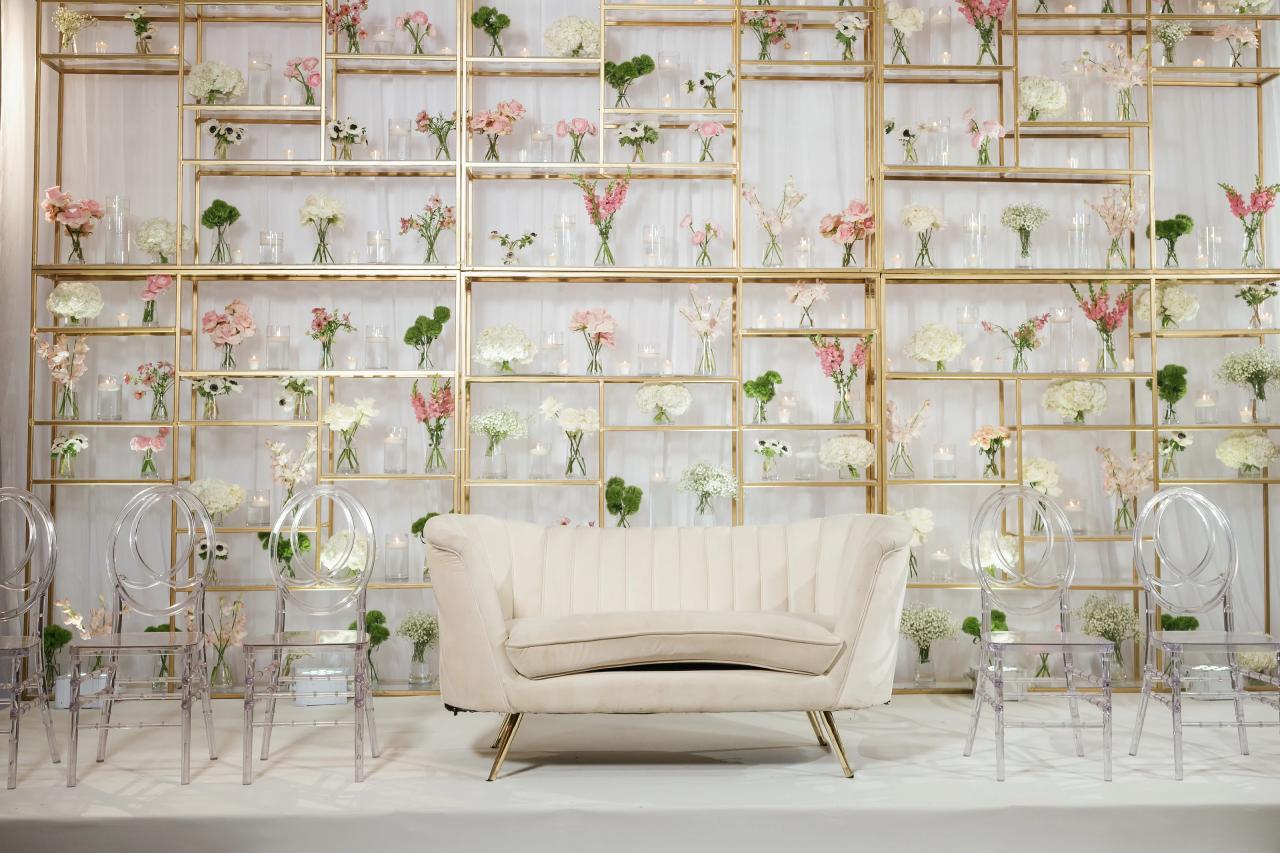 Elegant wedding stage with a white sofa centered before a gold geometric backdrop filled with pastel flowers and candle lights, flanked by clear acrylic chairs.