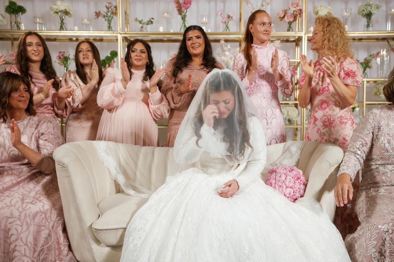 Bride seated in a white gown and veil with a hand near her face, looking down, as family members around her clap and smile in a softly lit floral ceremony space.