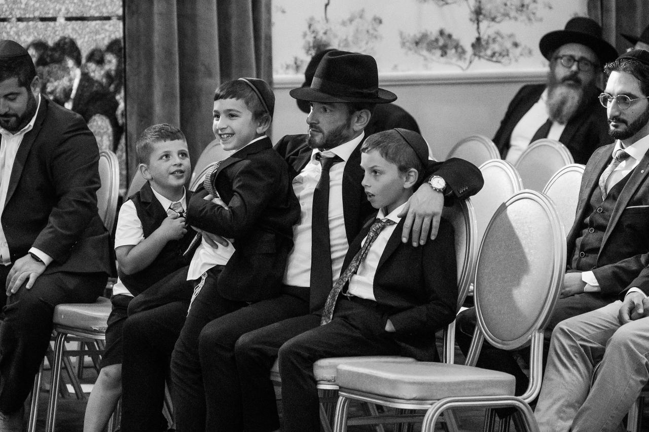 Black-and-white photo of a formal Jewish wedding/mitzvah ceremony seating area with seated family members and children in suits and kippot, looking attentively toward the right.