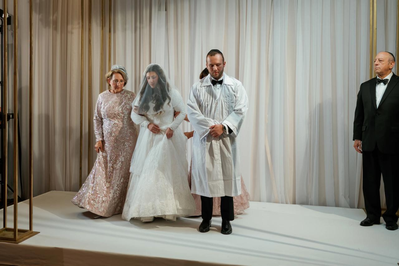 Bride and groom in white ritual attire walk forward indoors with family, framed against tall white drapes.