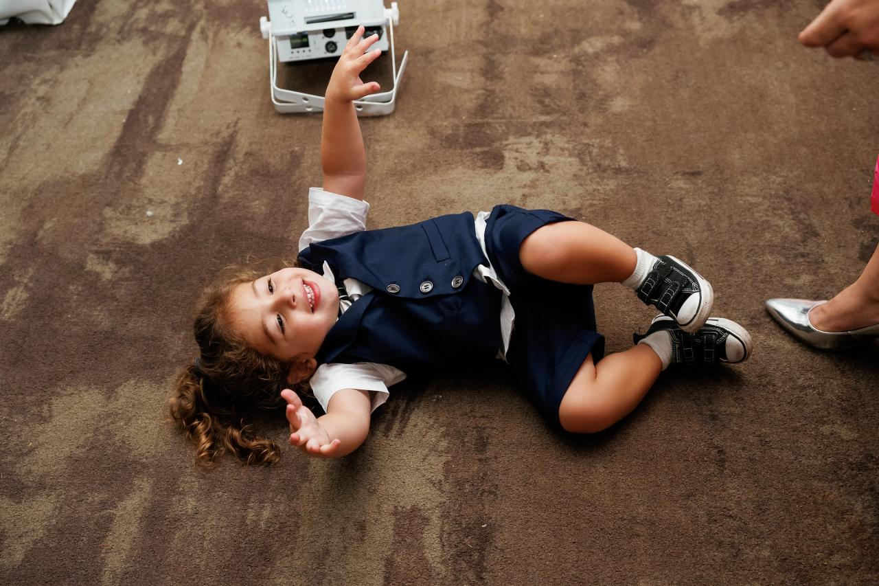 Child in a navy outfit lying on the floor with arms raised, looking up toward the camera at an indoor celebration.