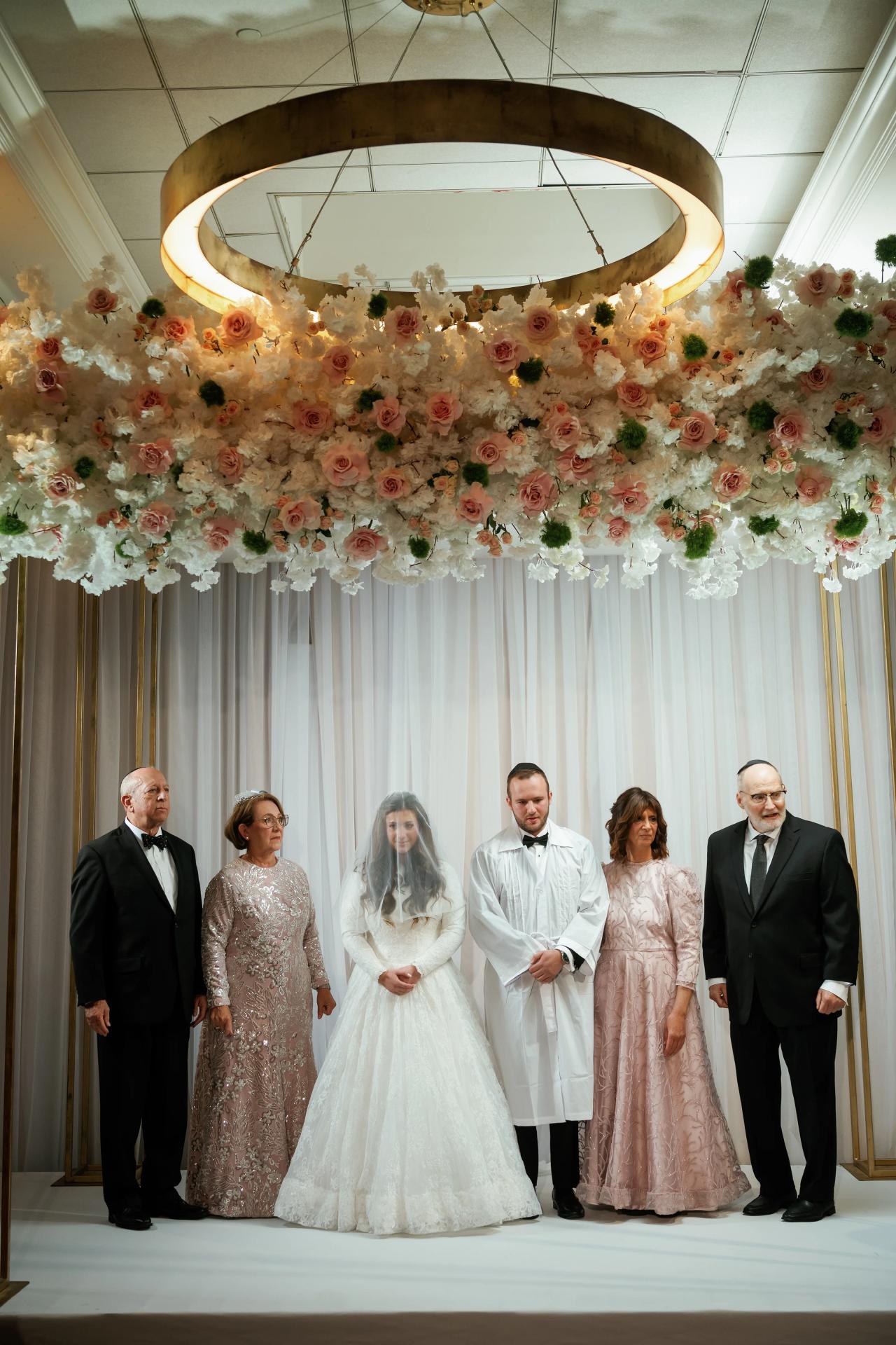 Group formal wedding portrait of the bride and groom centered with close family members standing beneath an elaborate overhead floral canopy with pink roses and greenery.