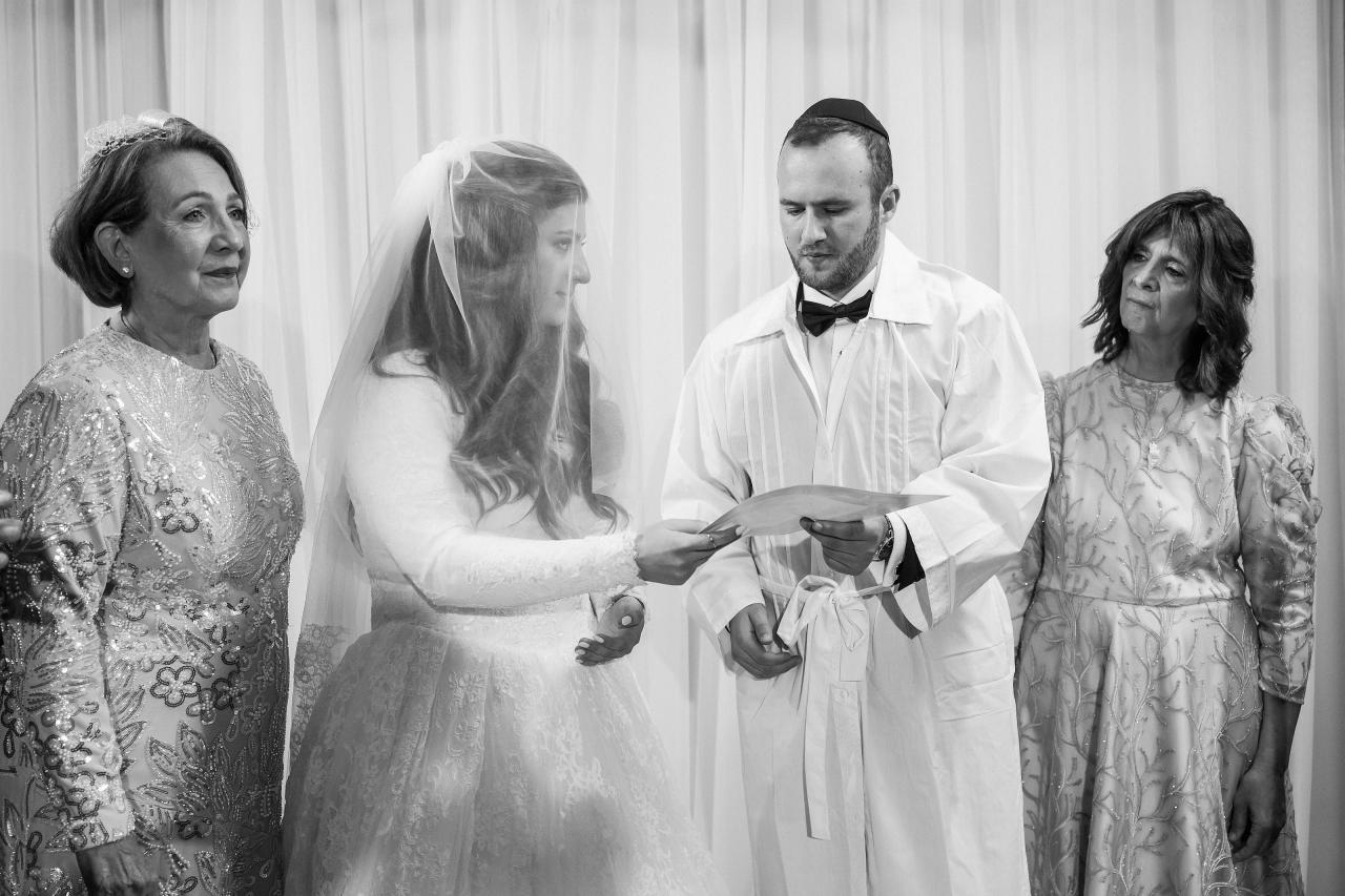 Black-and-white photo of a couple at a ritual moment, standing center with close family watching, the groom looking down at a held document and the bride holding an item at her hands.