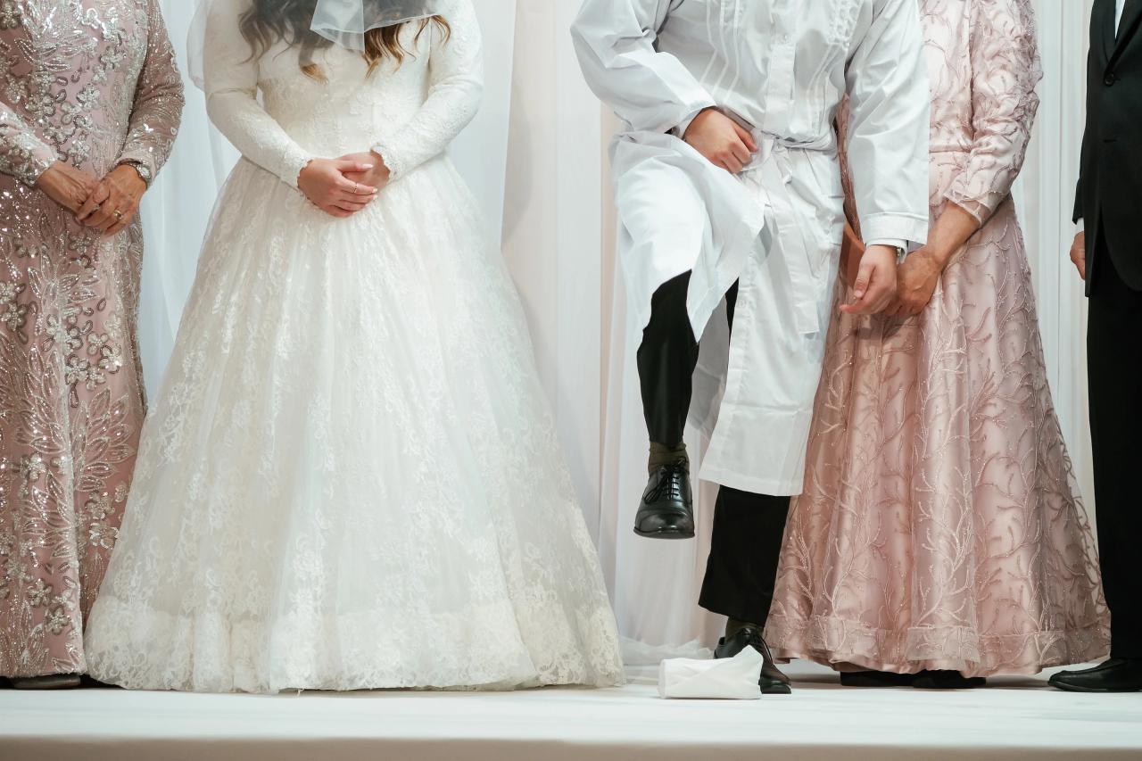 Close crop of a wedding ceremony moment showing the bride in a lace gown and the groom in a white outfit with hands held near the couple, with family attire visible at the edges.