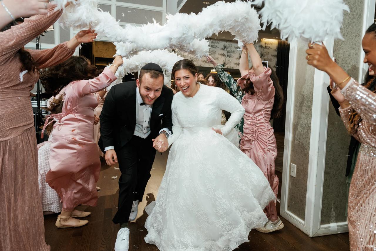 Bride and groom smiling during a lively reception celebration while guests around them cheer and raise feathered decorations