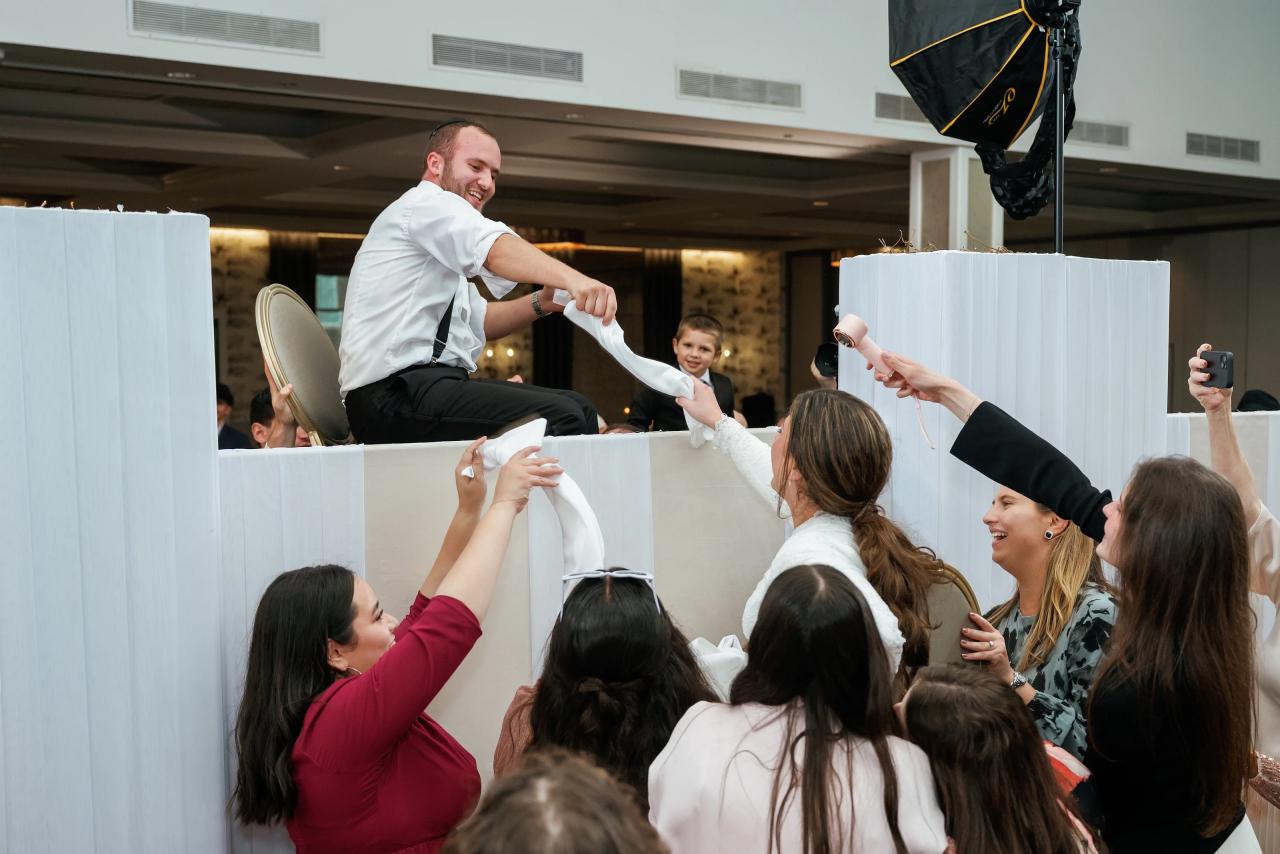 Smiling groom mid-jump above a white divider as guests reach upward cheering during the celebration.